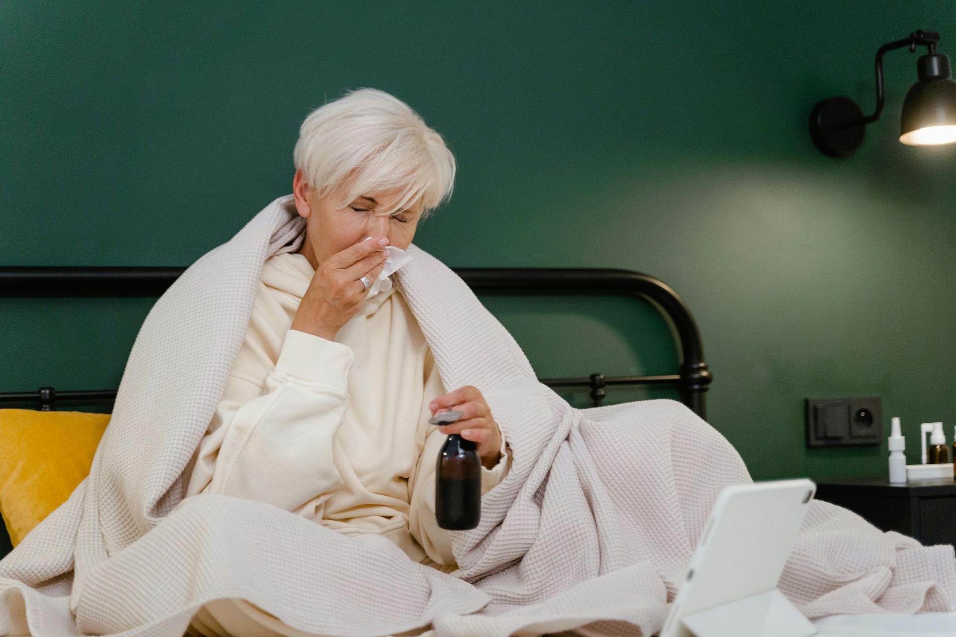 Woman in bed, blowing her nose, looking at a tablet, holding medicine. Covered in a blanket, green wall.