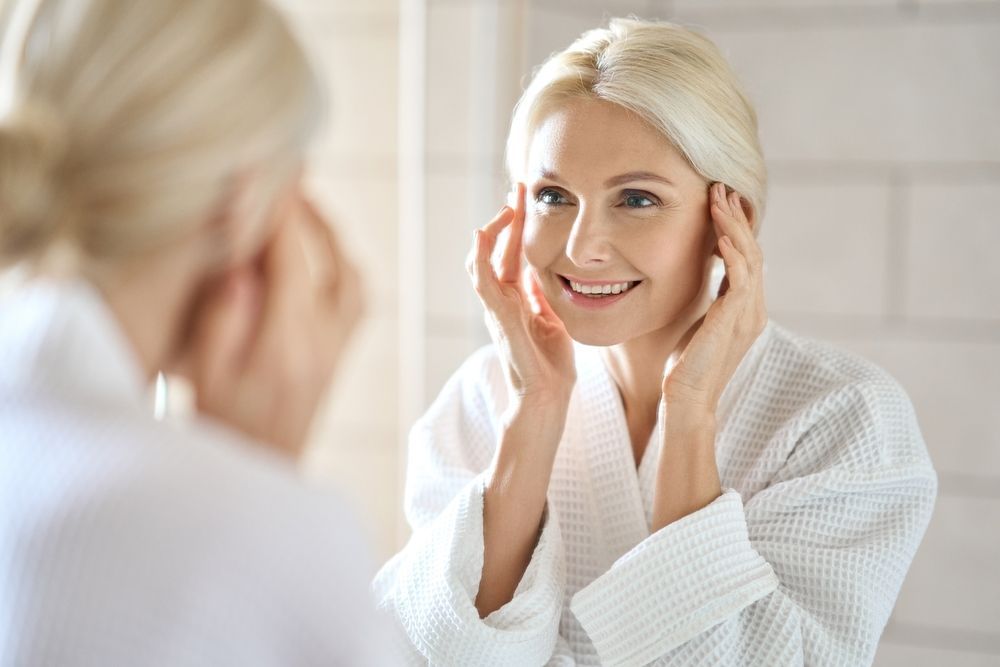 A woman in a bathrobe is looking at her face in the mirror.