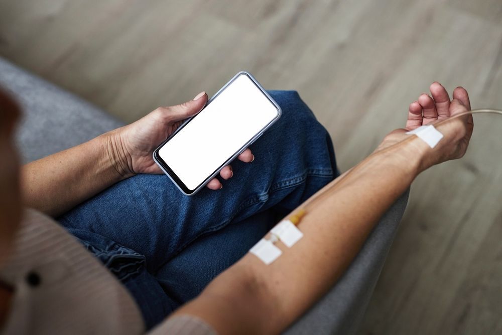 A woman is sitting on a couch holding a cell phone with a syringe in her arm.