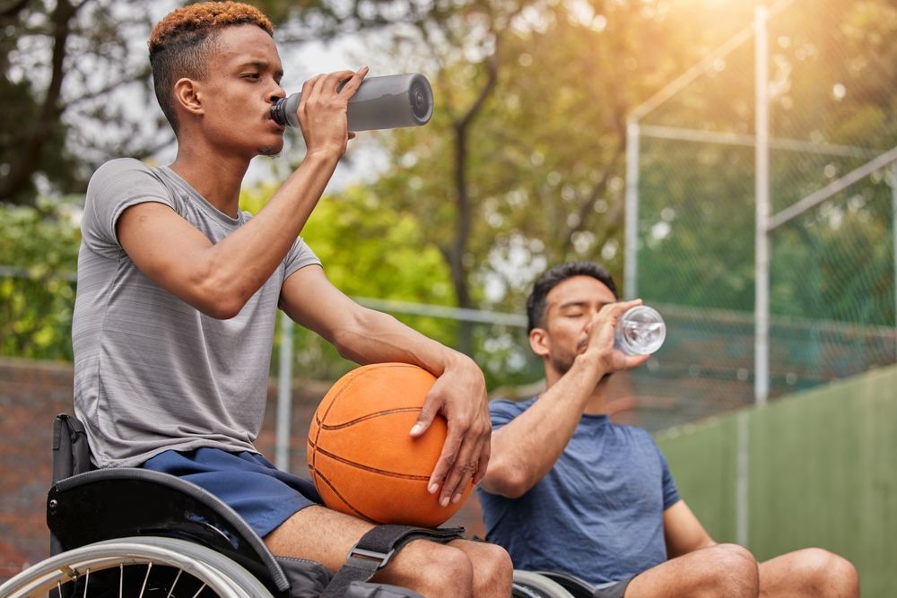 A man in a wheelchair is drinking water while another man holds a basketball.