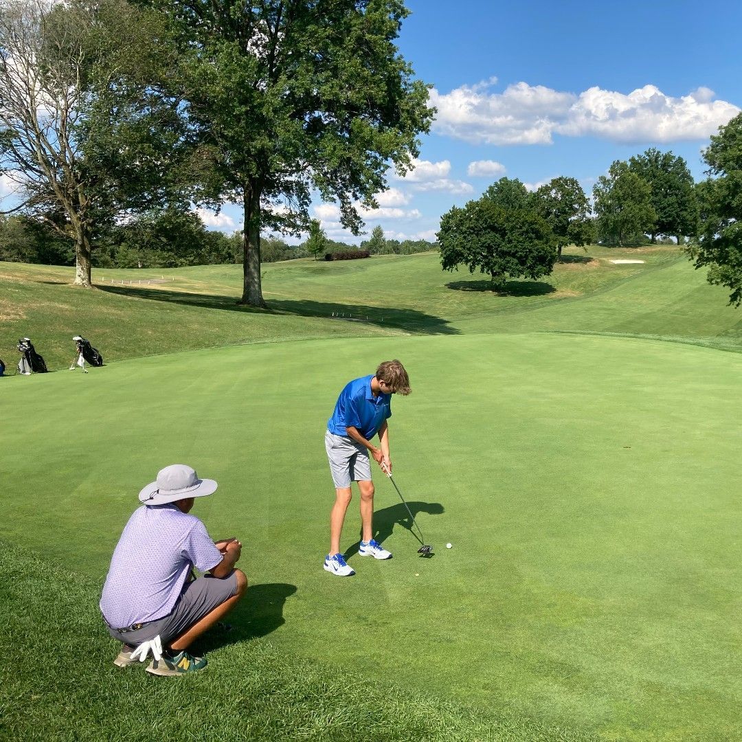 A person in a blue shirt putts on a golf green while an instructor in a sun hat crouches nearby to watch.