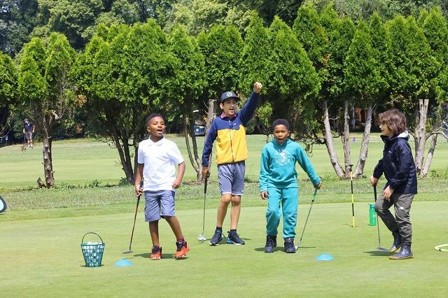 Four children standing on a green golf course, some holding clubs and posing joyfully near a practice bucket.