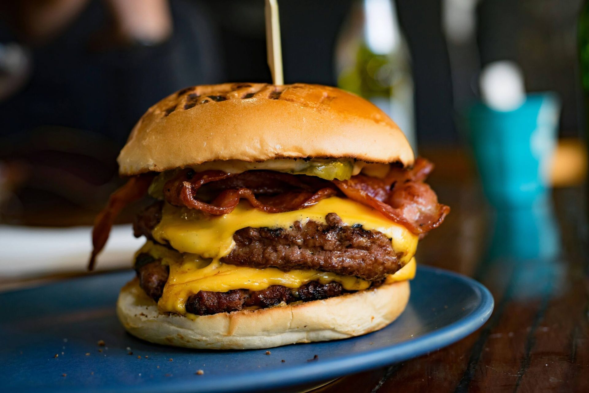 A double cheeseburger with bacon on a brioche bun sits on a blue plate at a table.