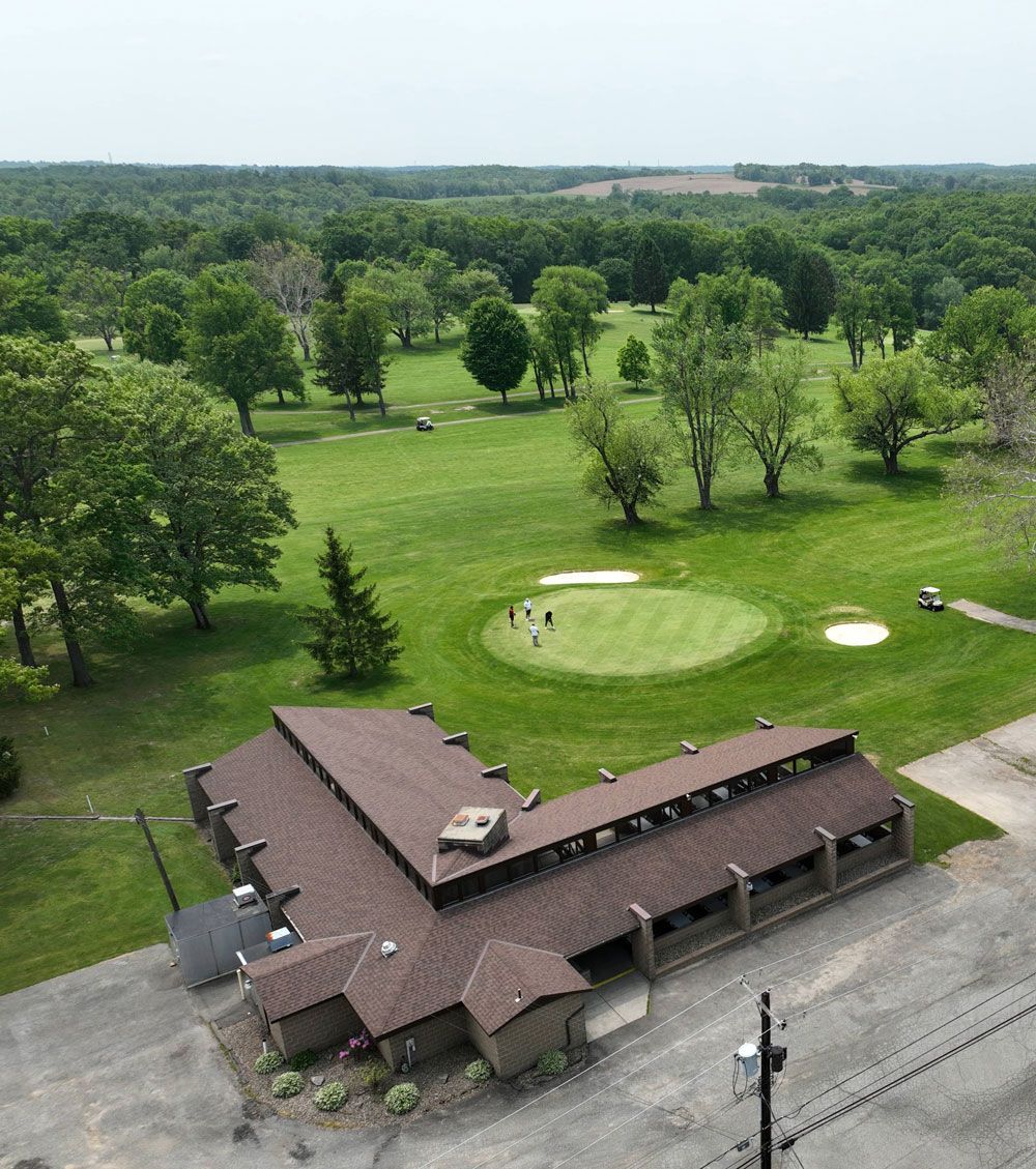 An aerial view of a building with a brown roof next to a putting green on a golf course surrounded by trees.