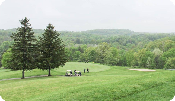 Golfers with carts on a lush green course, framed by two tall pine trees, set against a backdrop of rolling forest hills.