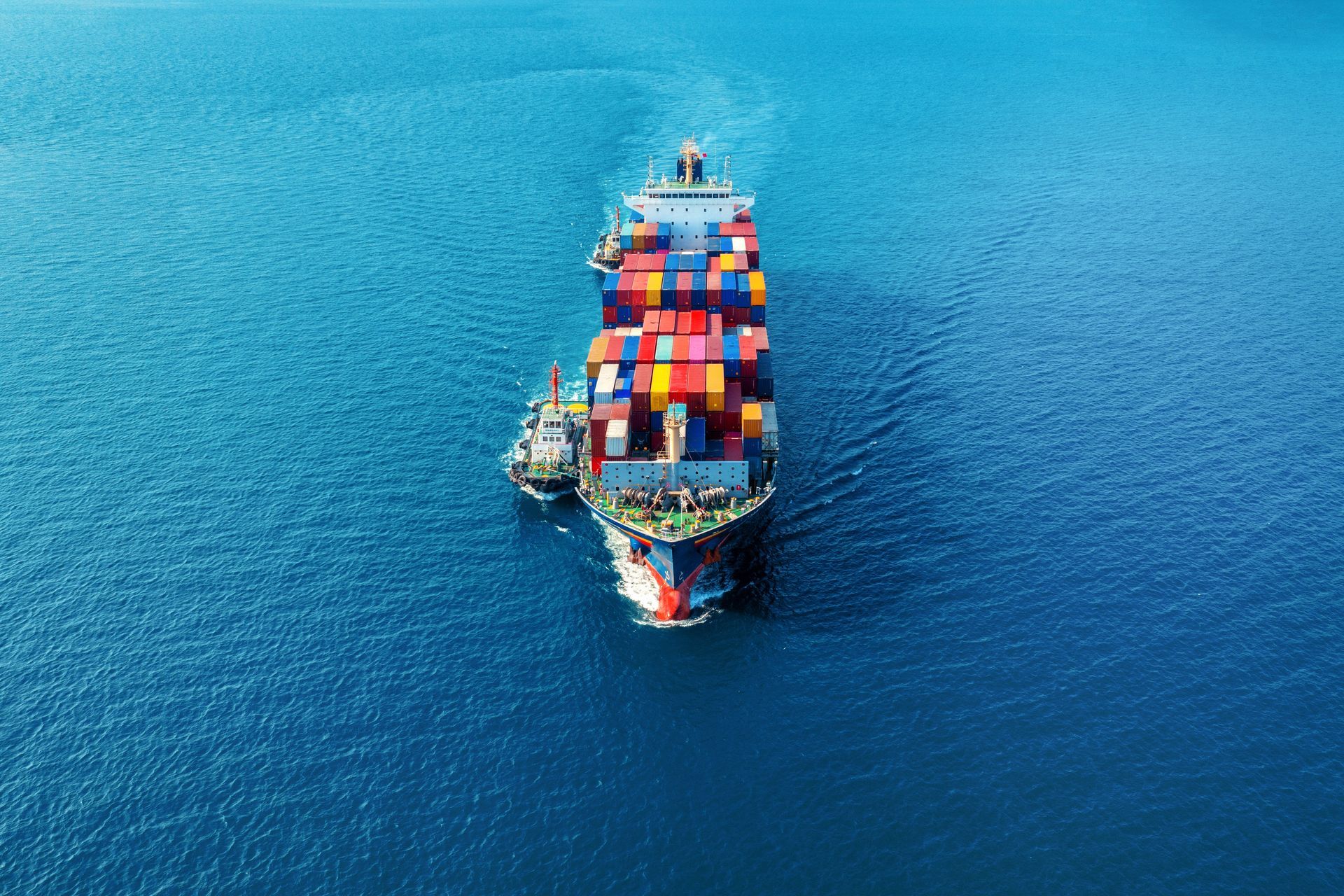 An aerial view of a colorful container ship being escorted by a smaller tugboat on open blue ocean water.