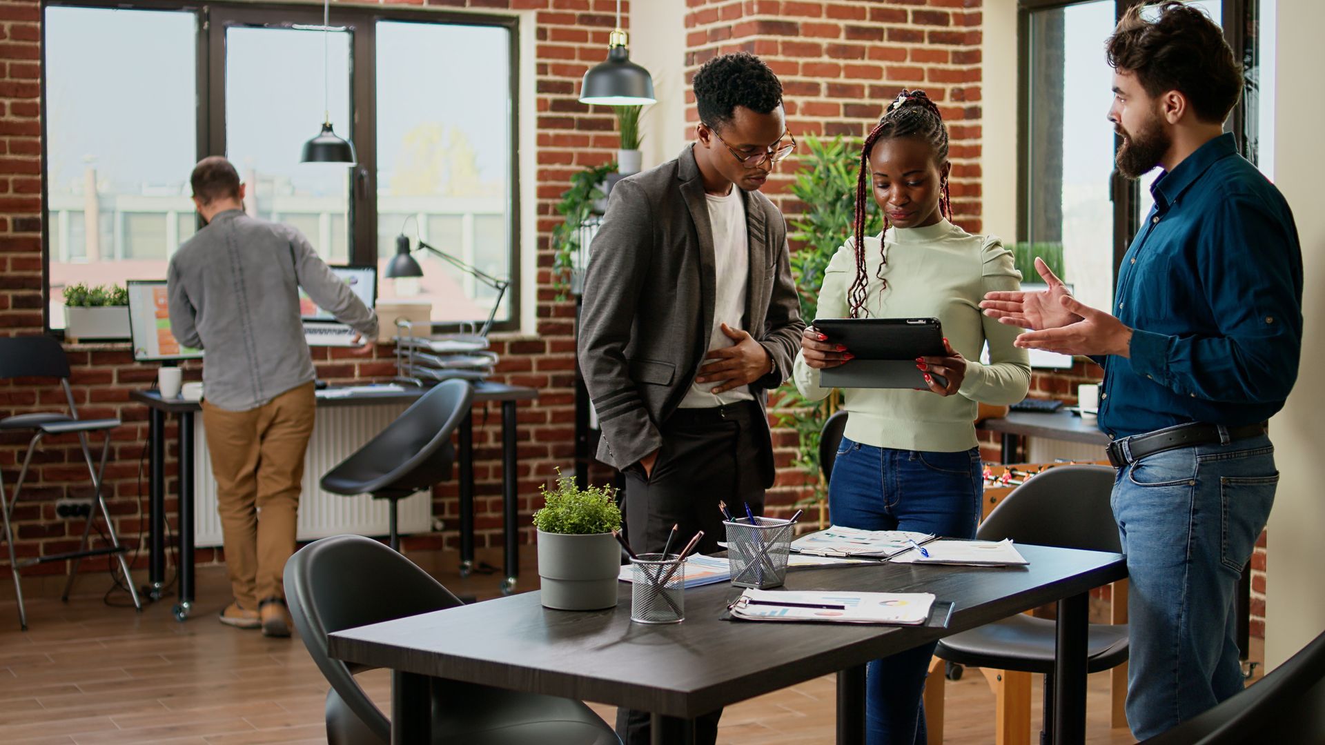 Three professionals collaborate in an office, one presenting on a tablet while another stands in the background by a desk.