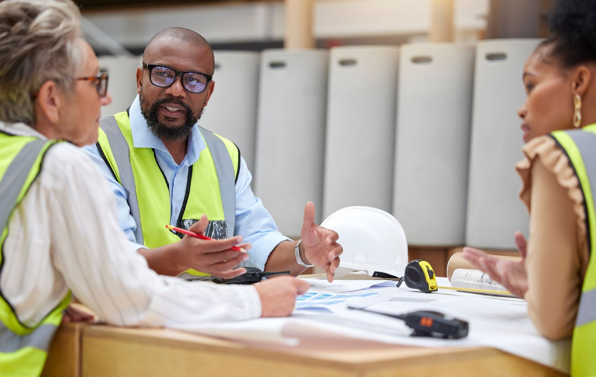 Three professionals in high-visibility vests collaborate over blueprints at a desk in a workshop setting.