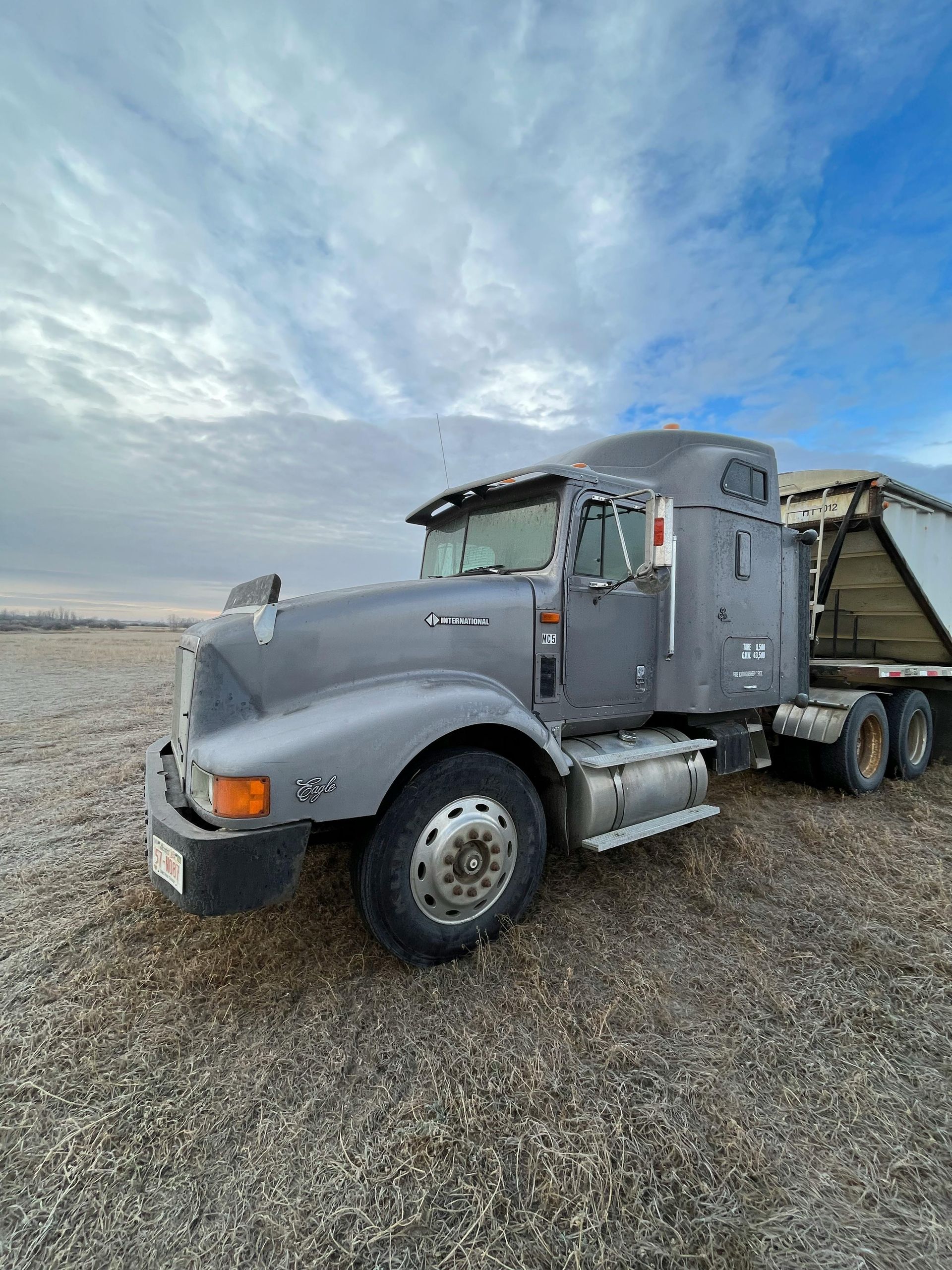 A gray semi-truck with a sleeper cab hitched to a trailer, parked on a gravel field under a cloudy sky.