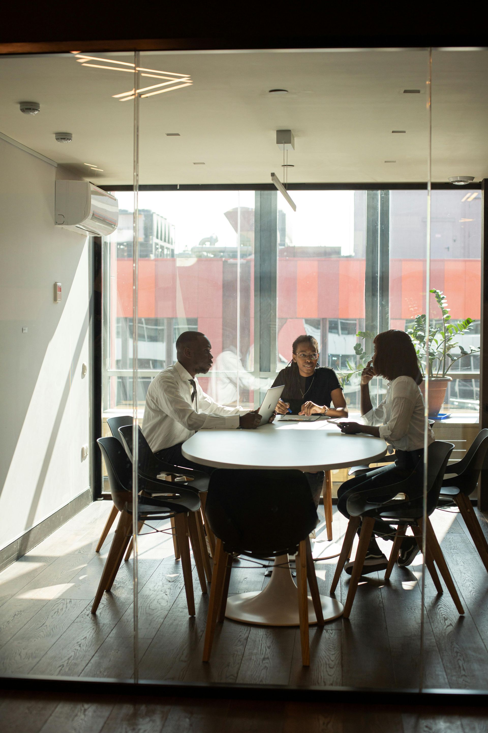 Three people sitting around a round table in a glass-walled conference room during a meeting.