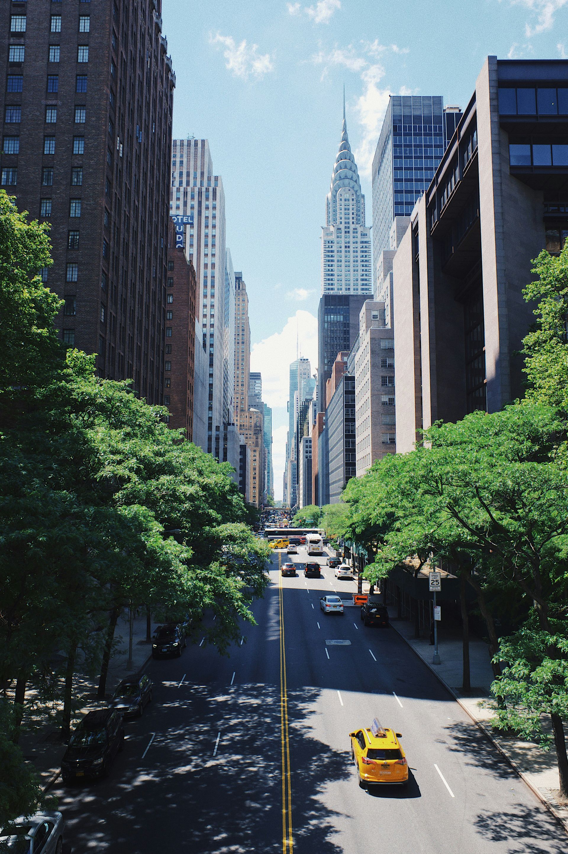 A yellow taxi drives down a New York City street flanked by trees and skyscrapers, including the Chrysler Building.
