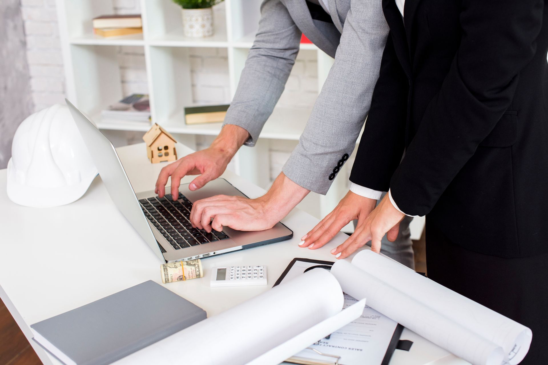 Two professionals collaborate at a desk with a laptop, architectural blueprints, a model house, and a white hard hat.