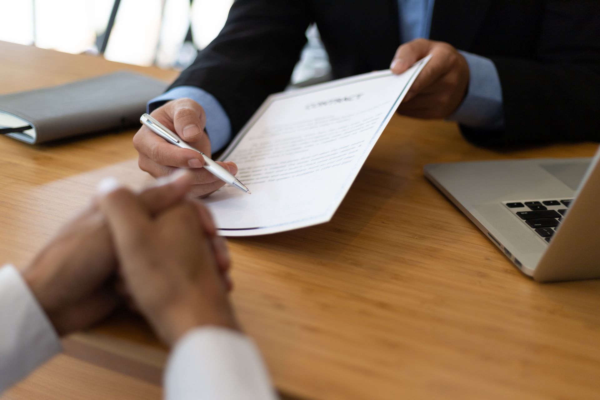 A person in a suit hands a contract and a pen to another person sitting across a wooden desk with a laptop.