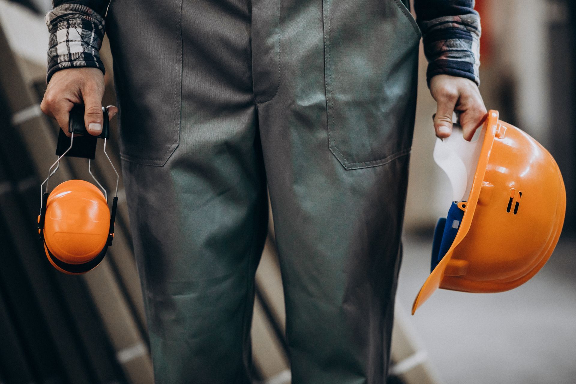 A worker wearing gray work pants holds orange earmuffs in one hand and an orange hard hat in the other.