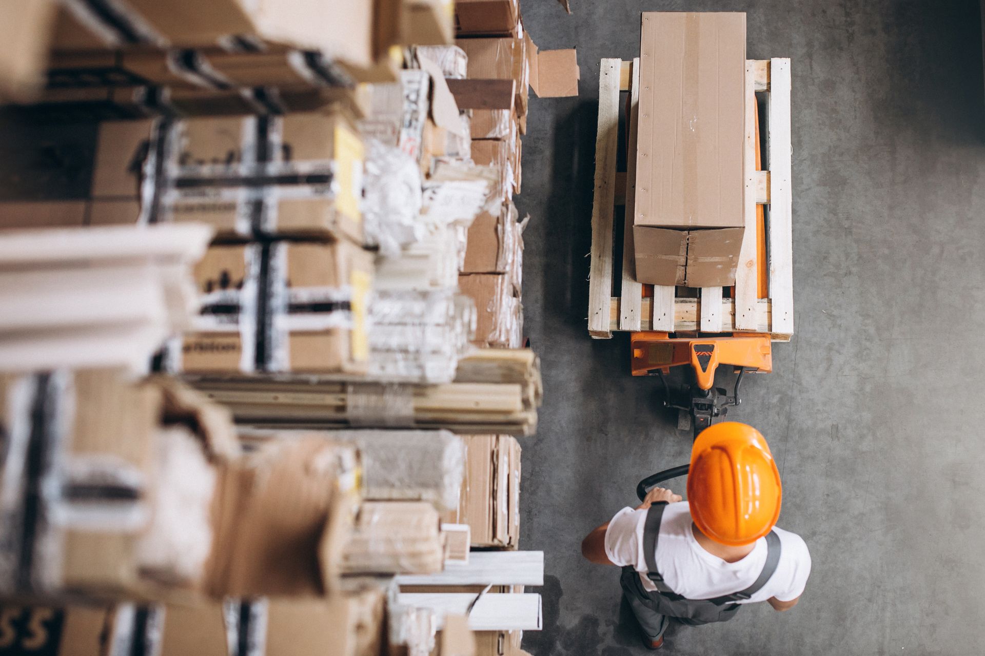 An overhead view of a worker in a hard hat moving a cardboard box on a wooden pallet with a manual pallet jack.