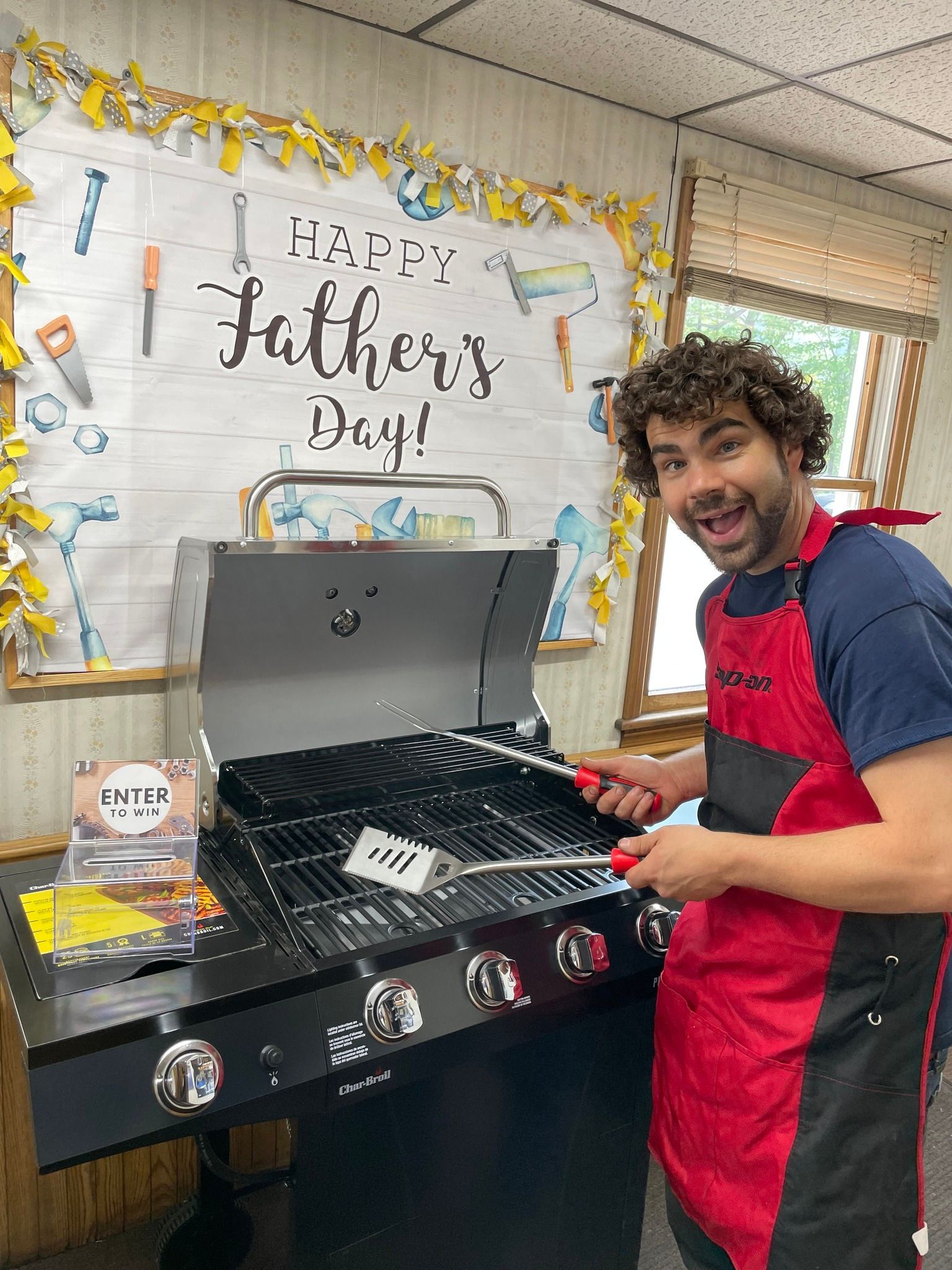 Man in red apron smiles, holding grilling tools over new grill. Happy Father's Day banner in background. | Forward Motors