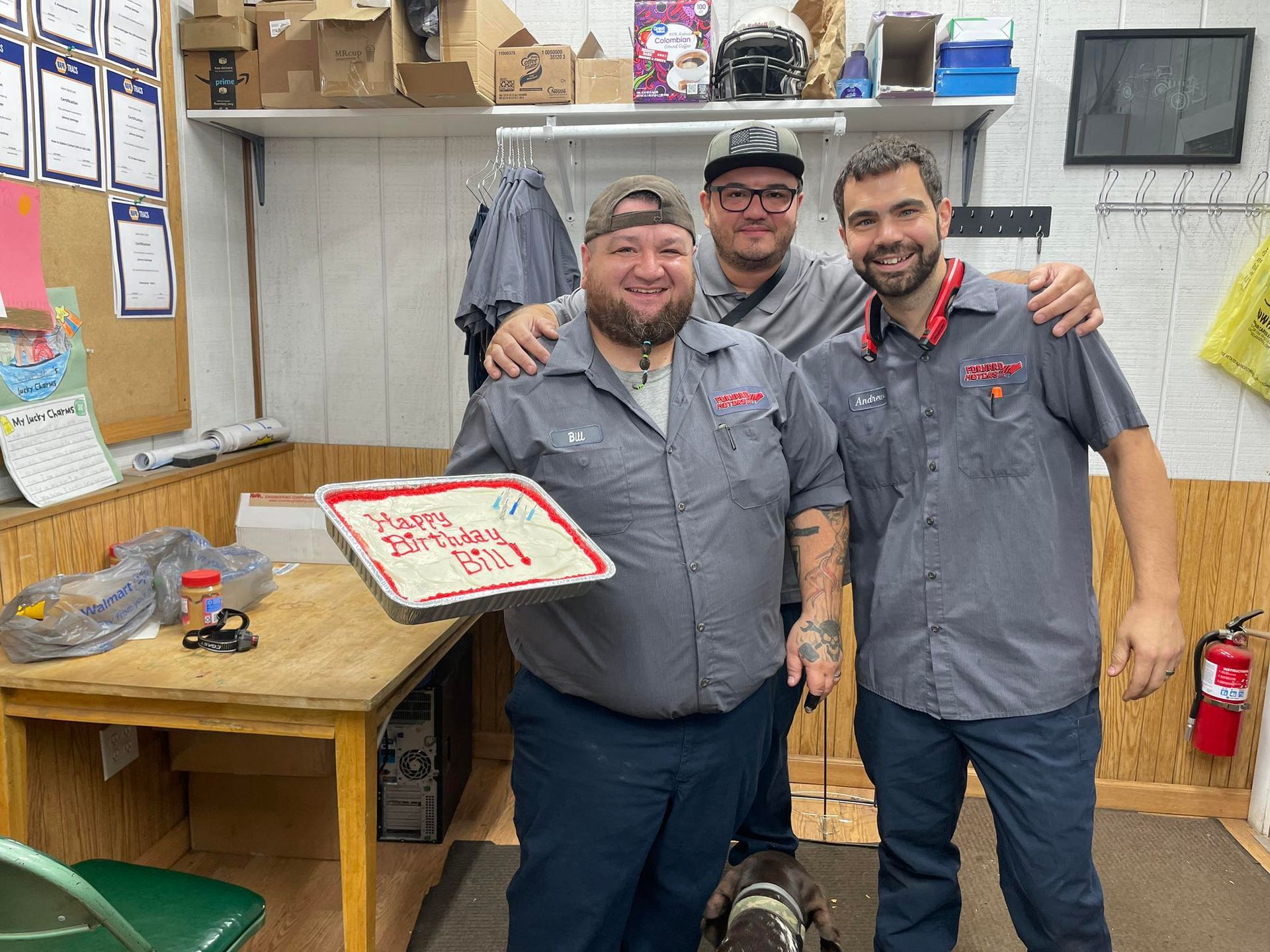 Three men in work uniforms celebrate a birthday with cake indoors.