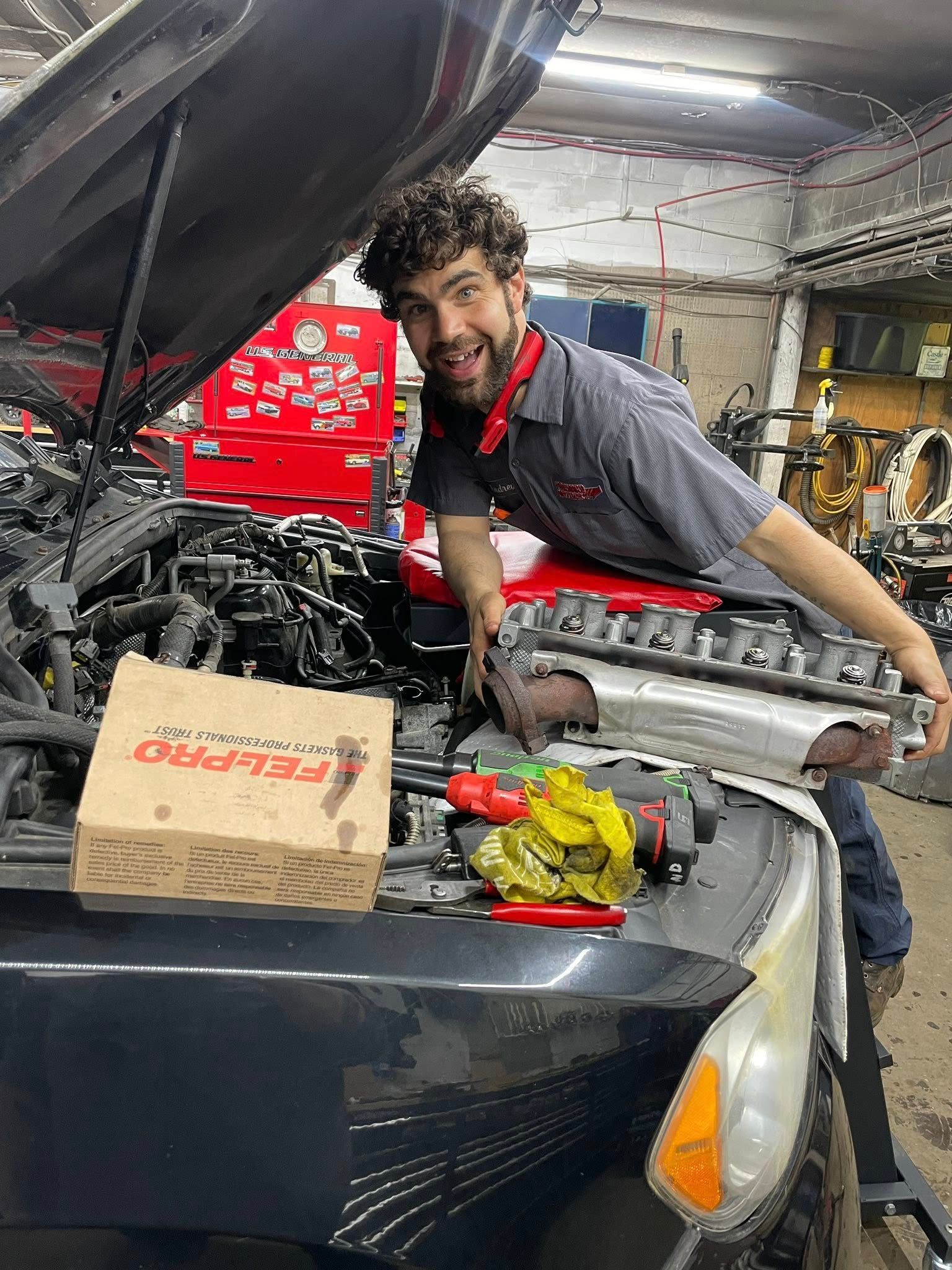 Mechanic with curly hair holds car engine part, smiling in a garage. Car hood open, tool box in background.
