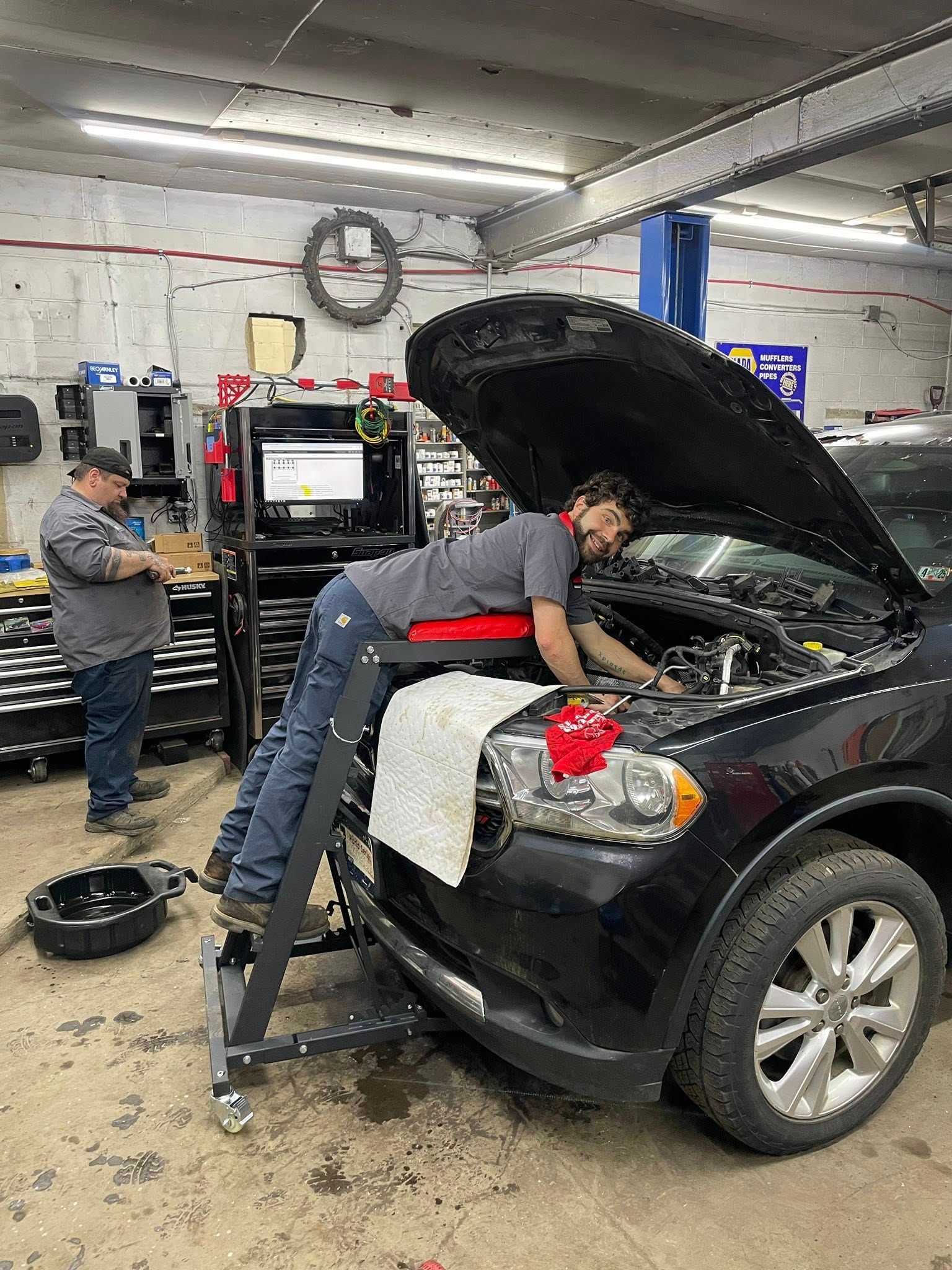 Mechanic working on a car engine. Another mechanic looks on, tools and equipment visible.