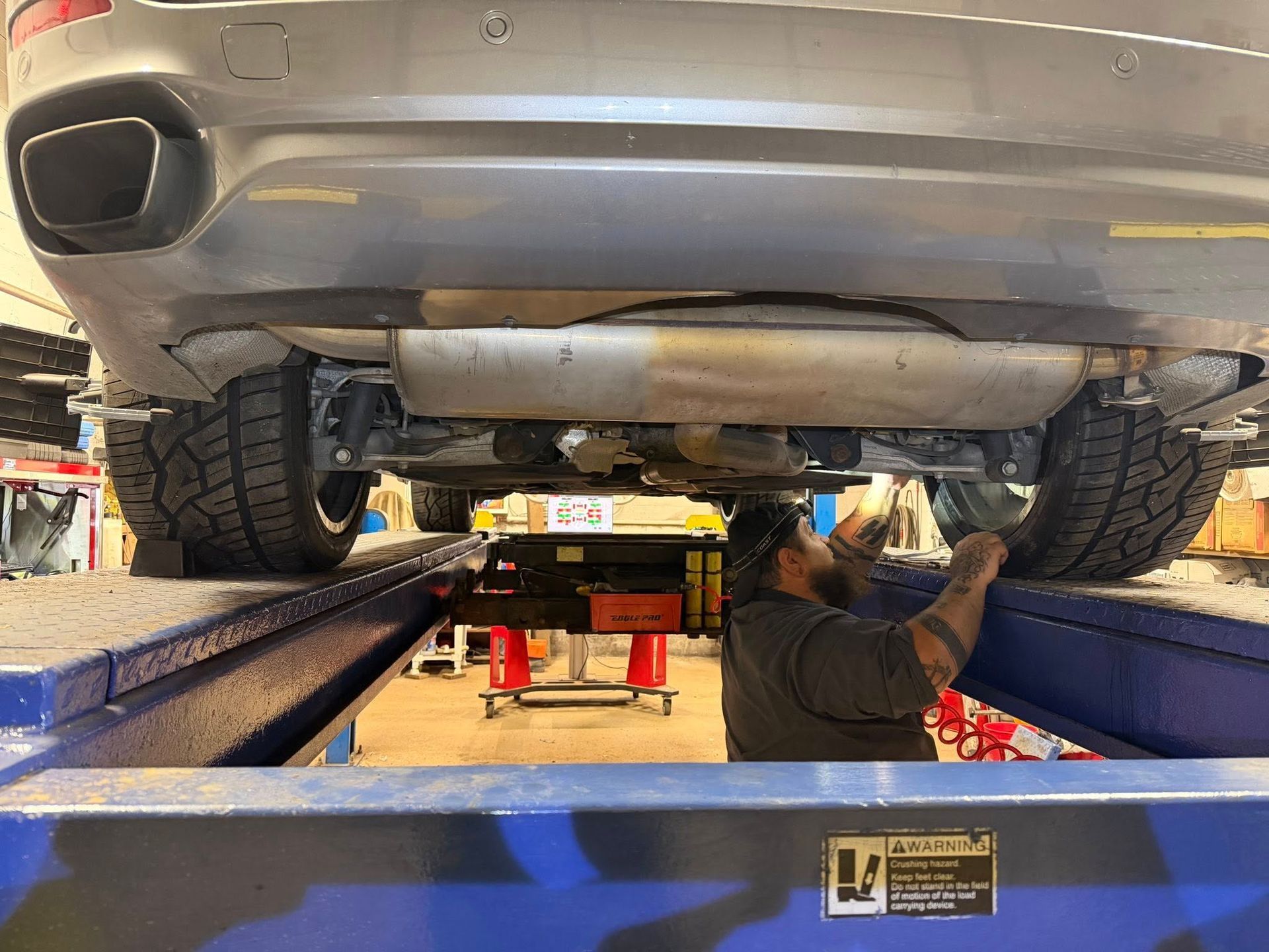 A mechanic working under a gray car raised on a blue lift in a garage.