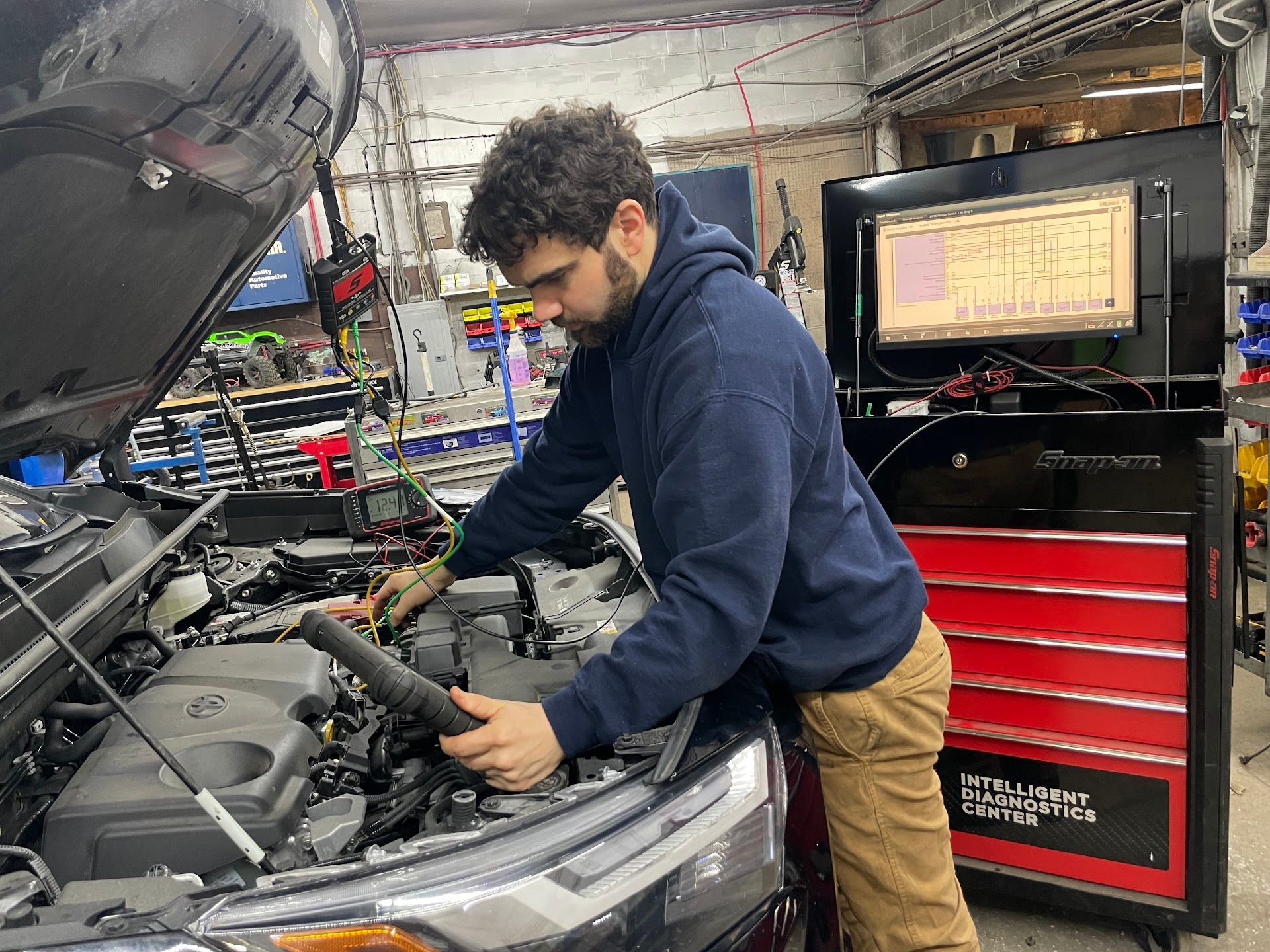 Mechanic working on car engine, diagnostic tool visible. Dark hair, blue hoodie, tan pants in a shop.