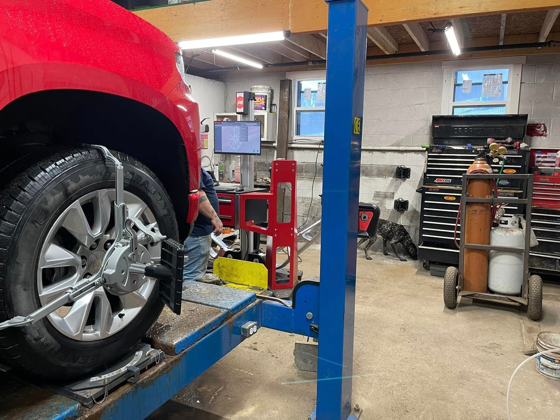 Red truck on a car lift in a garage undergoing wheel alignment. Tools and equipment visible.