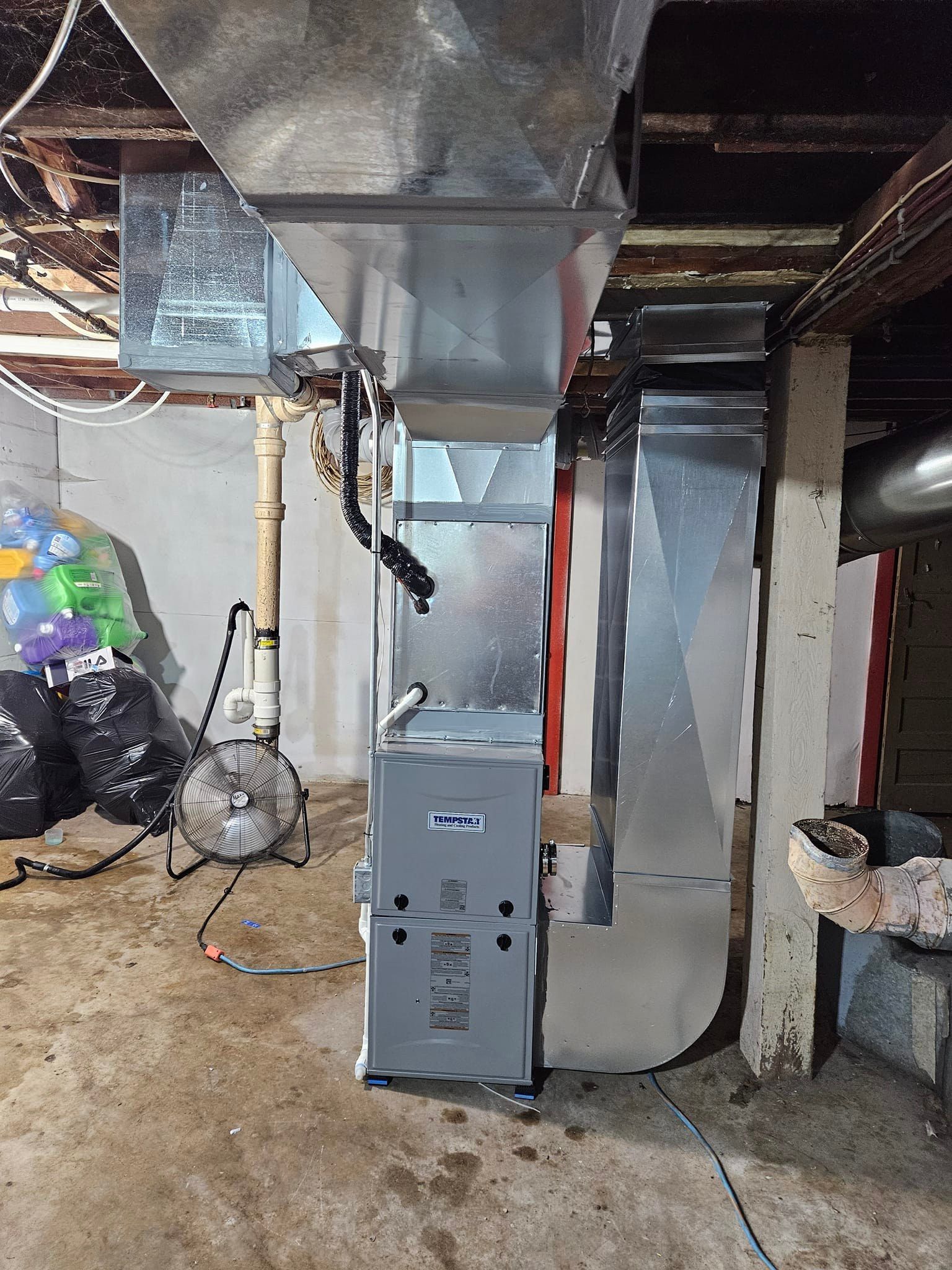 A furnace is sitting in a basement next to a fan.