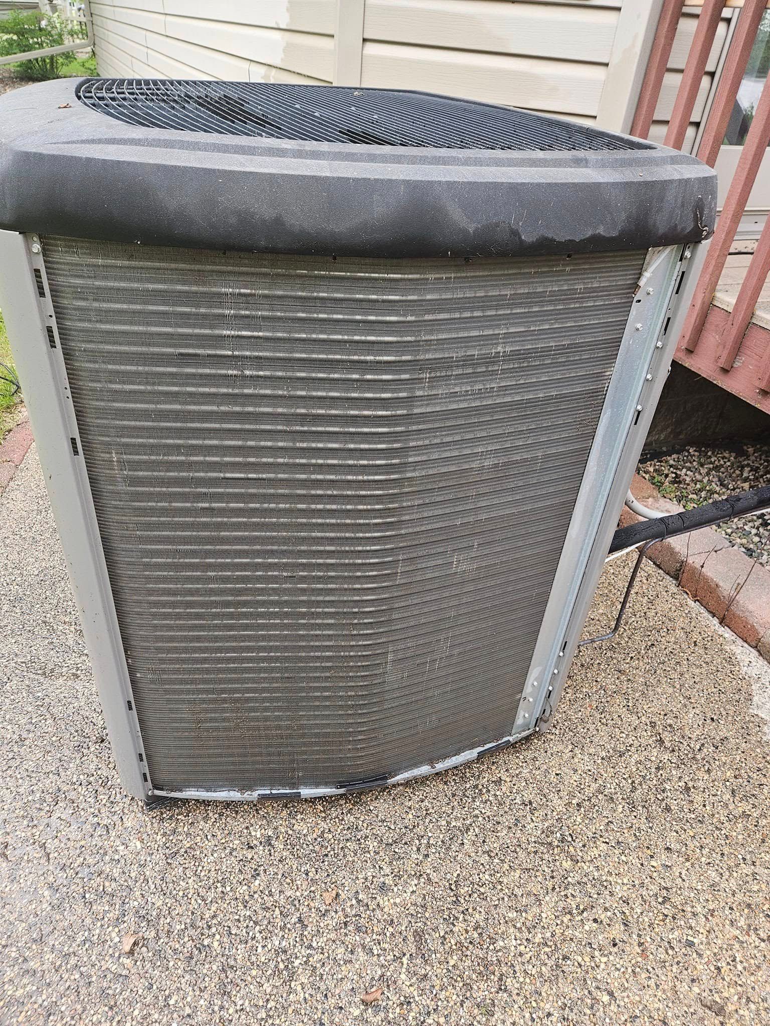 A dirty air conditioner is sitting on the sidewalk next to a house.