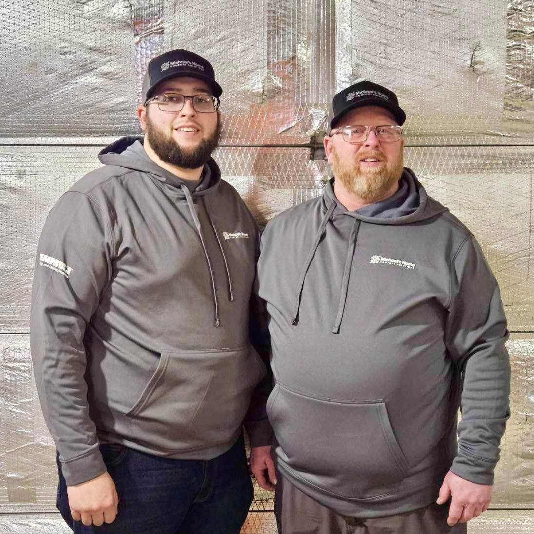 Two men are standing next to each other in front of a wooden wall.