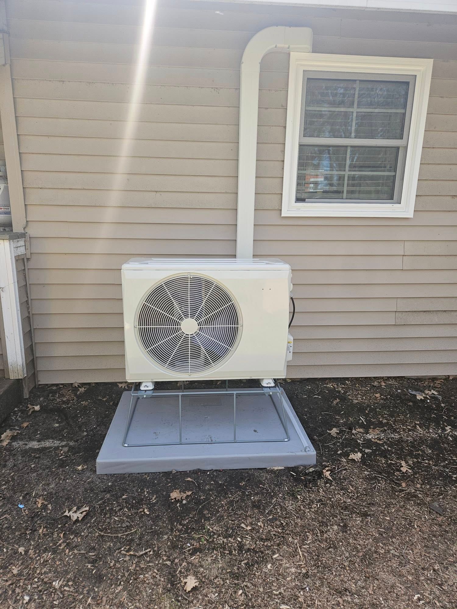 A white air conditioner is mounted on the side of a house next to a window.