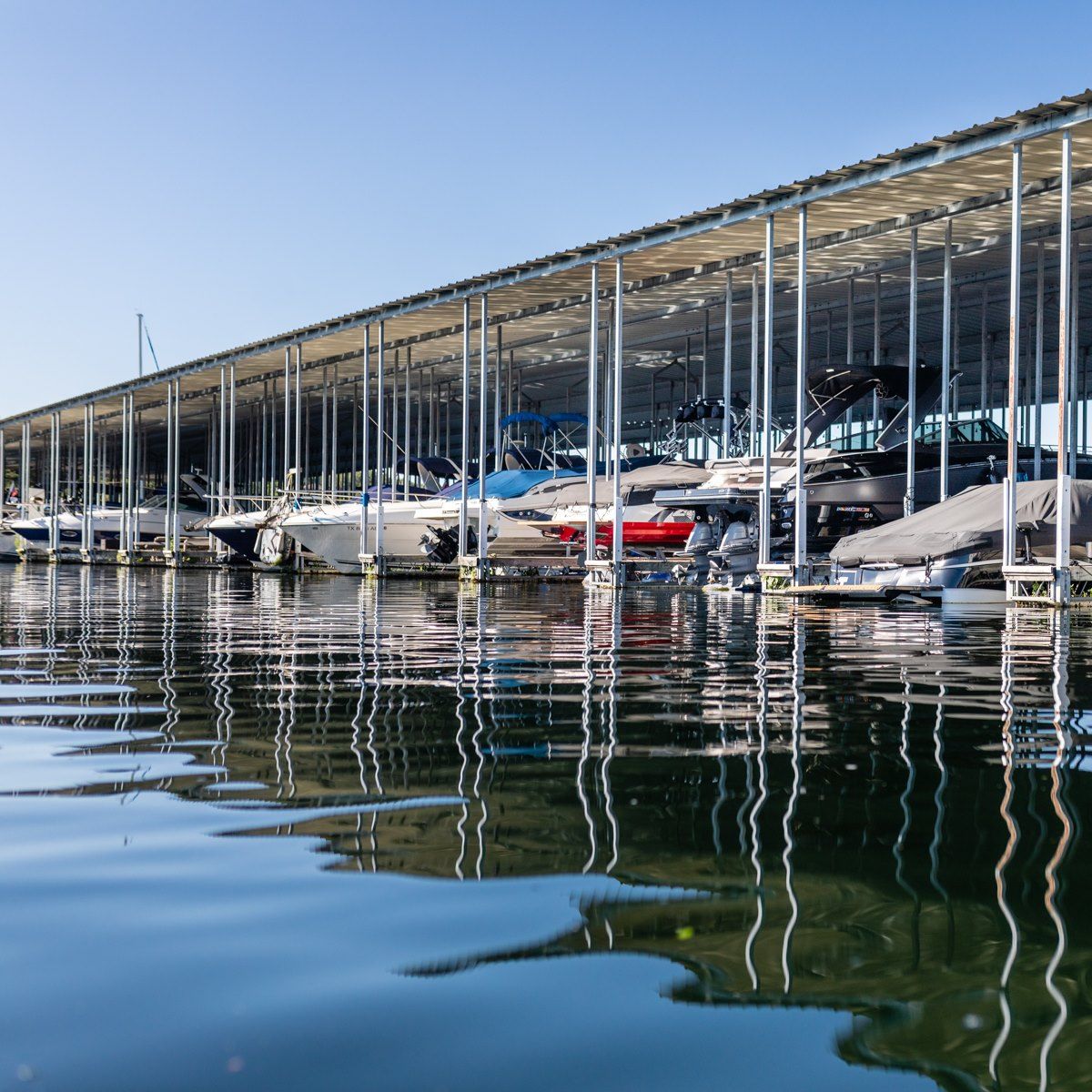 Boats docked under a long covered shelter, reflected in the water, on a sunny day.