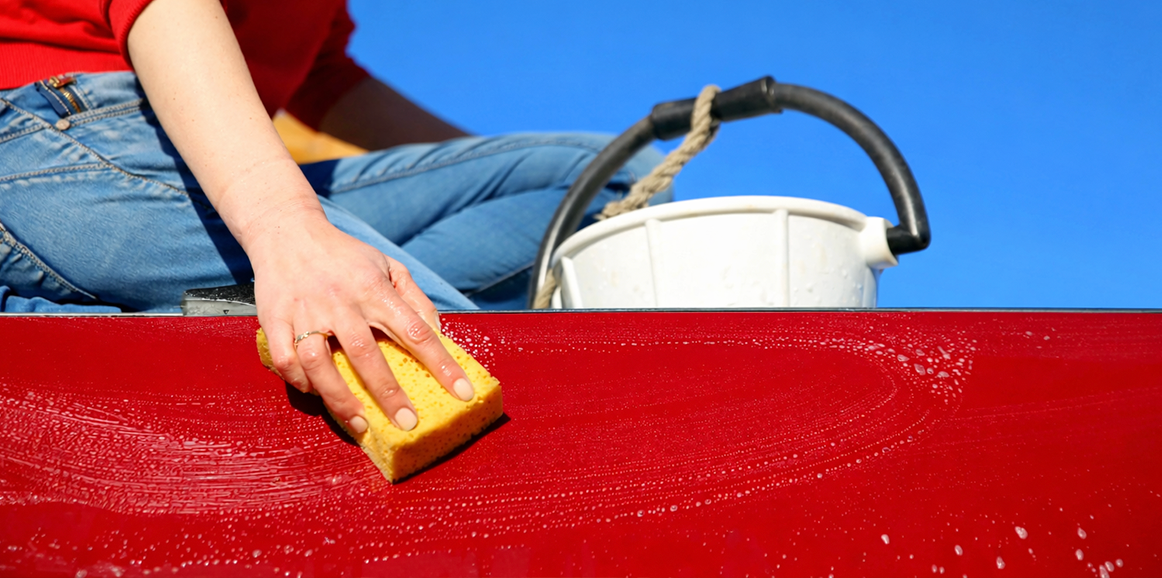 A person uses a sponge to wipe a red surface; a white bucket sits nearby, blue sky in background.