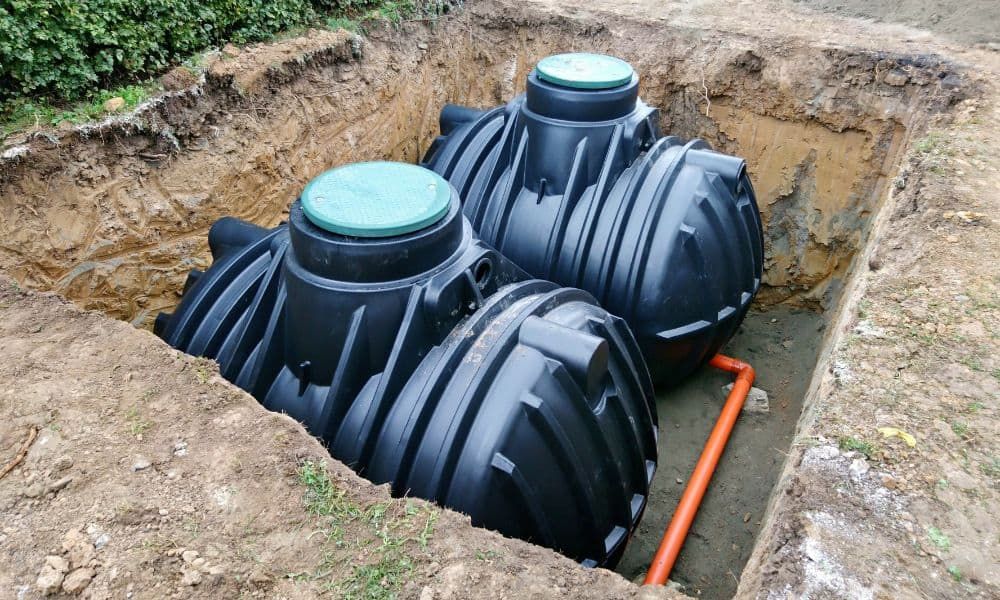 Two black plastic septic tanks with green lids installed in a rectangular dirt excavation.