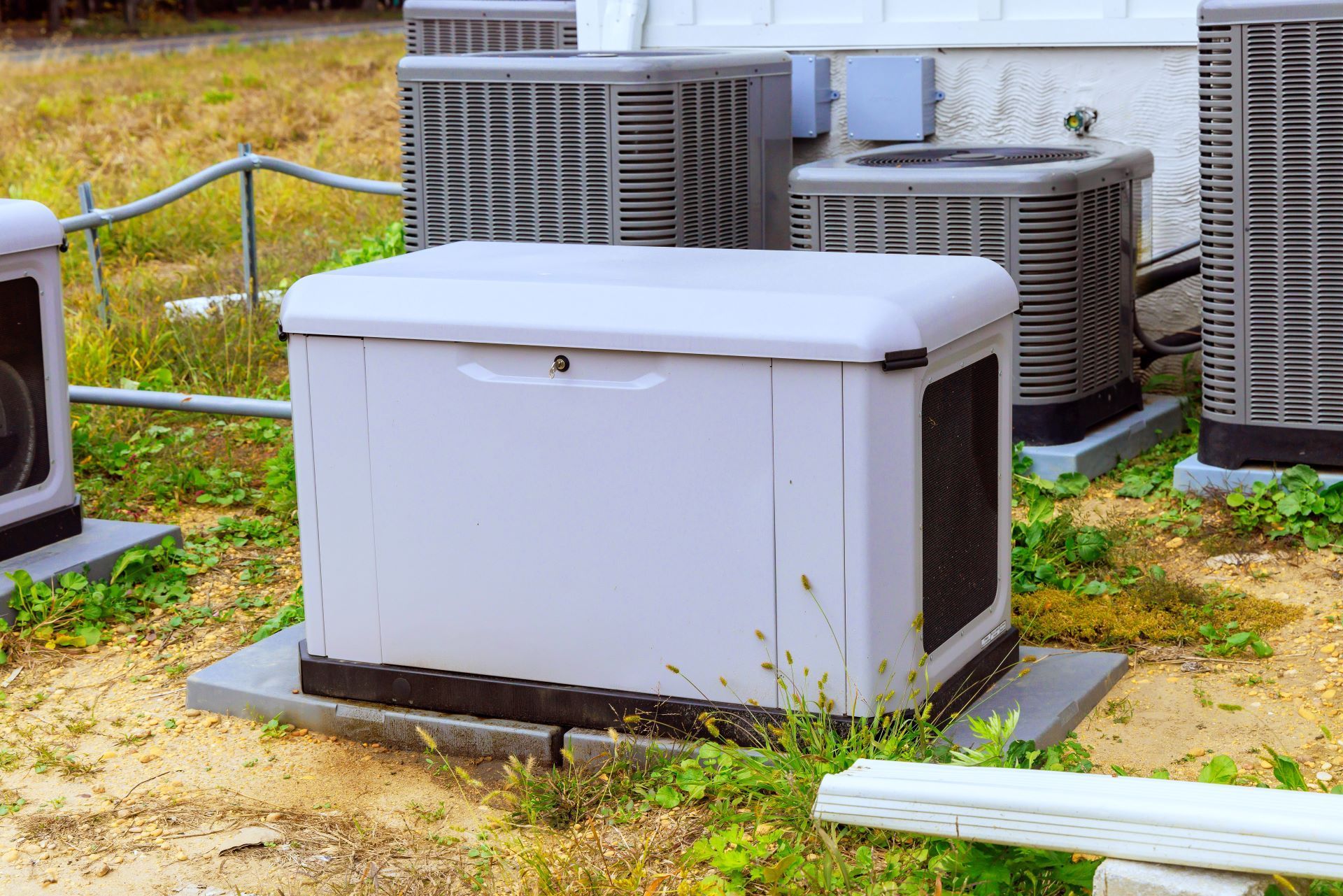 A gray standby generator surrounded by air conditioning units on a gravel patch.
