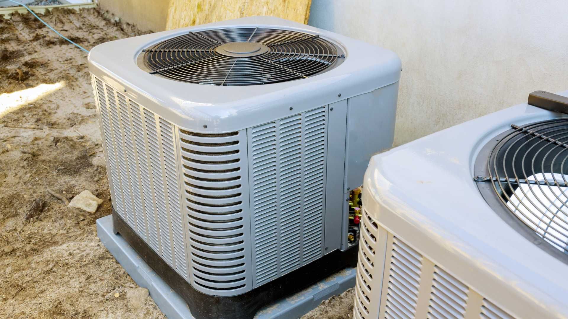 Two air conditioners are sitting next to each other in the dirt.