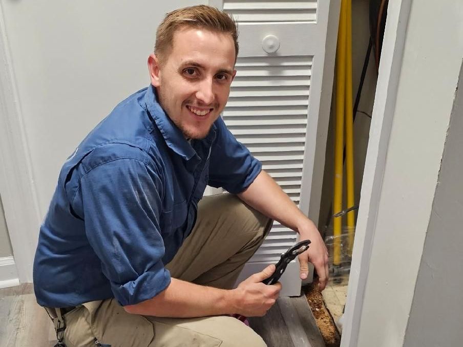 A man in a blue shirt is kneeling down in a closet holding a tool.
