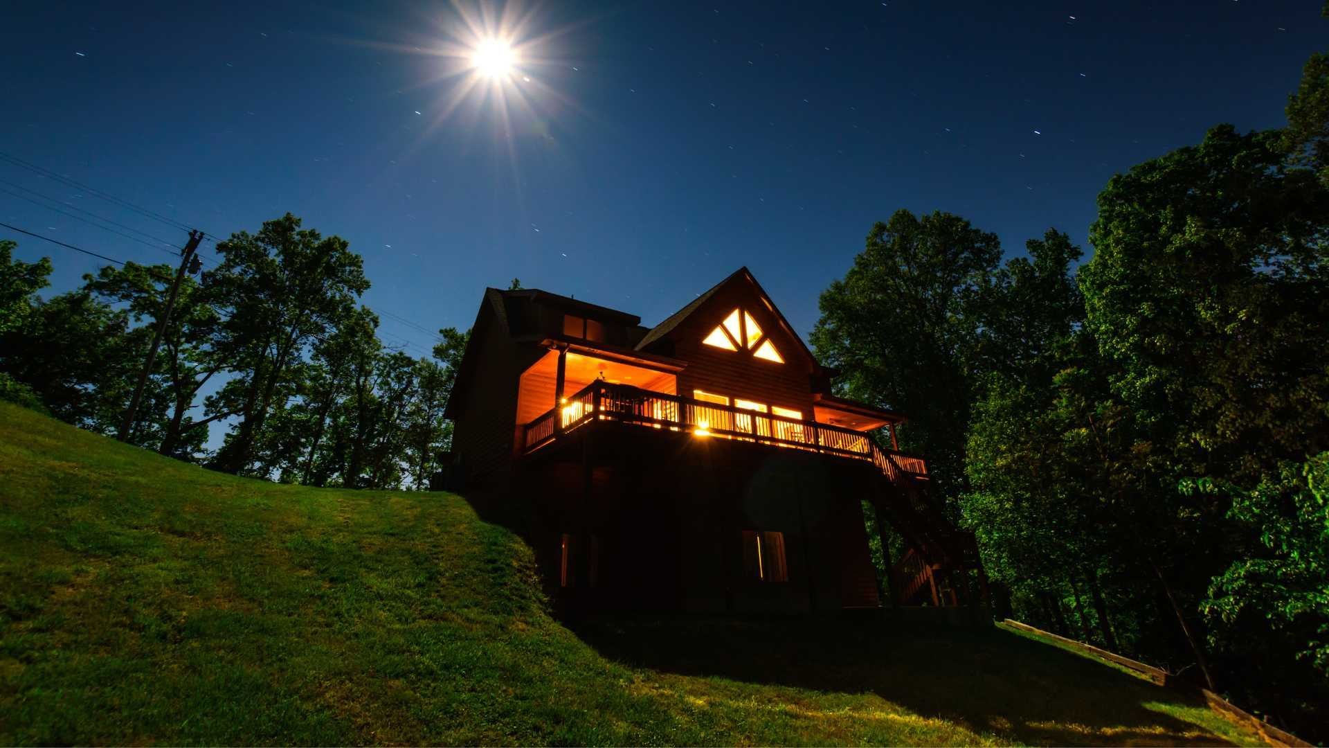 A cabin is lit up at night with a full moon in the sky.