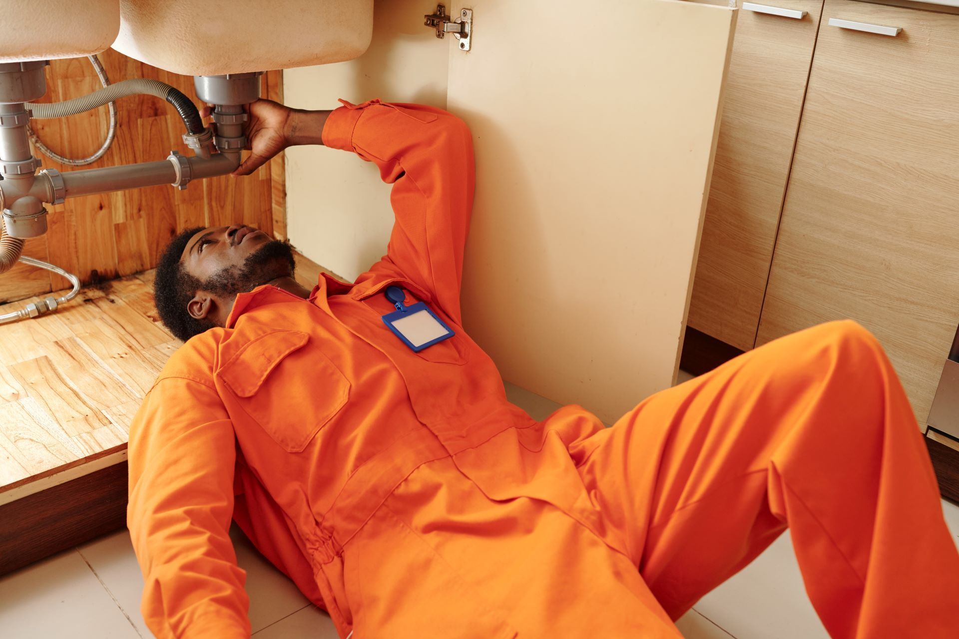A person in orange coveralls is lying on the floor under a sink, working on the plumbing.