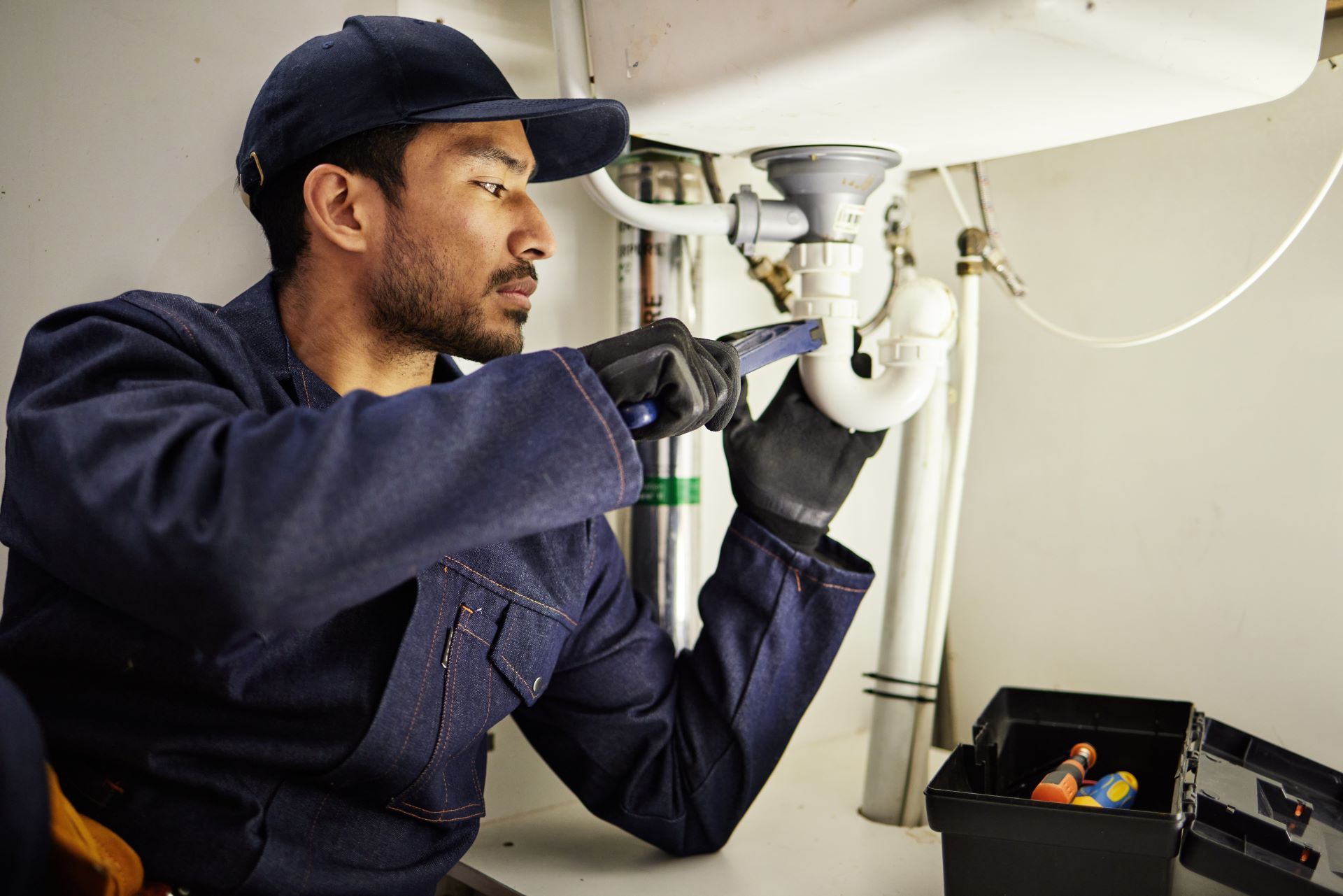 A plumber wearing a cap and work gloves repairs a white P-trap pipe under a sink.