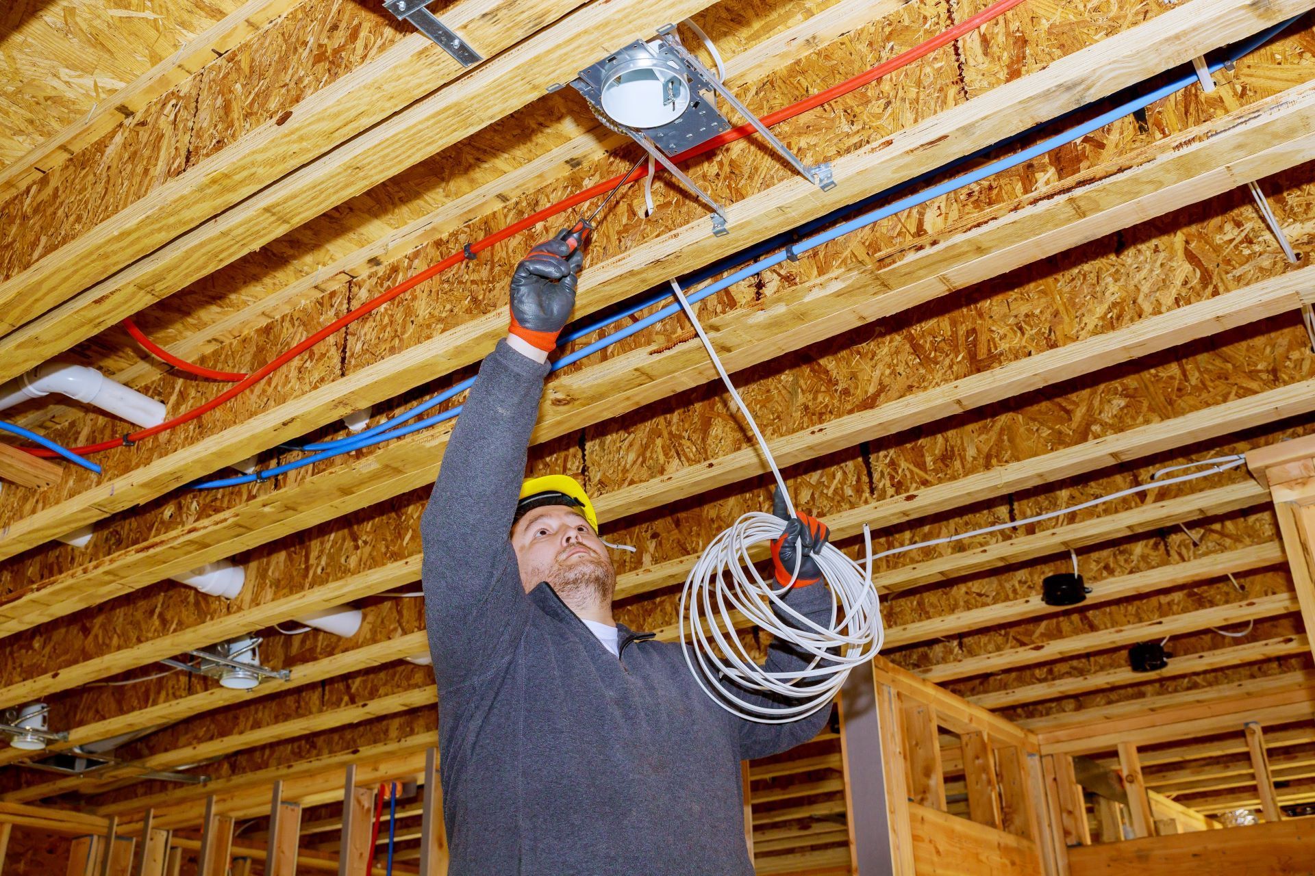 An electrician wearing a hard hat installs electrical wiring in the exposed wooden ceiling of a house under construction.