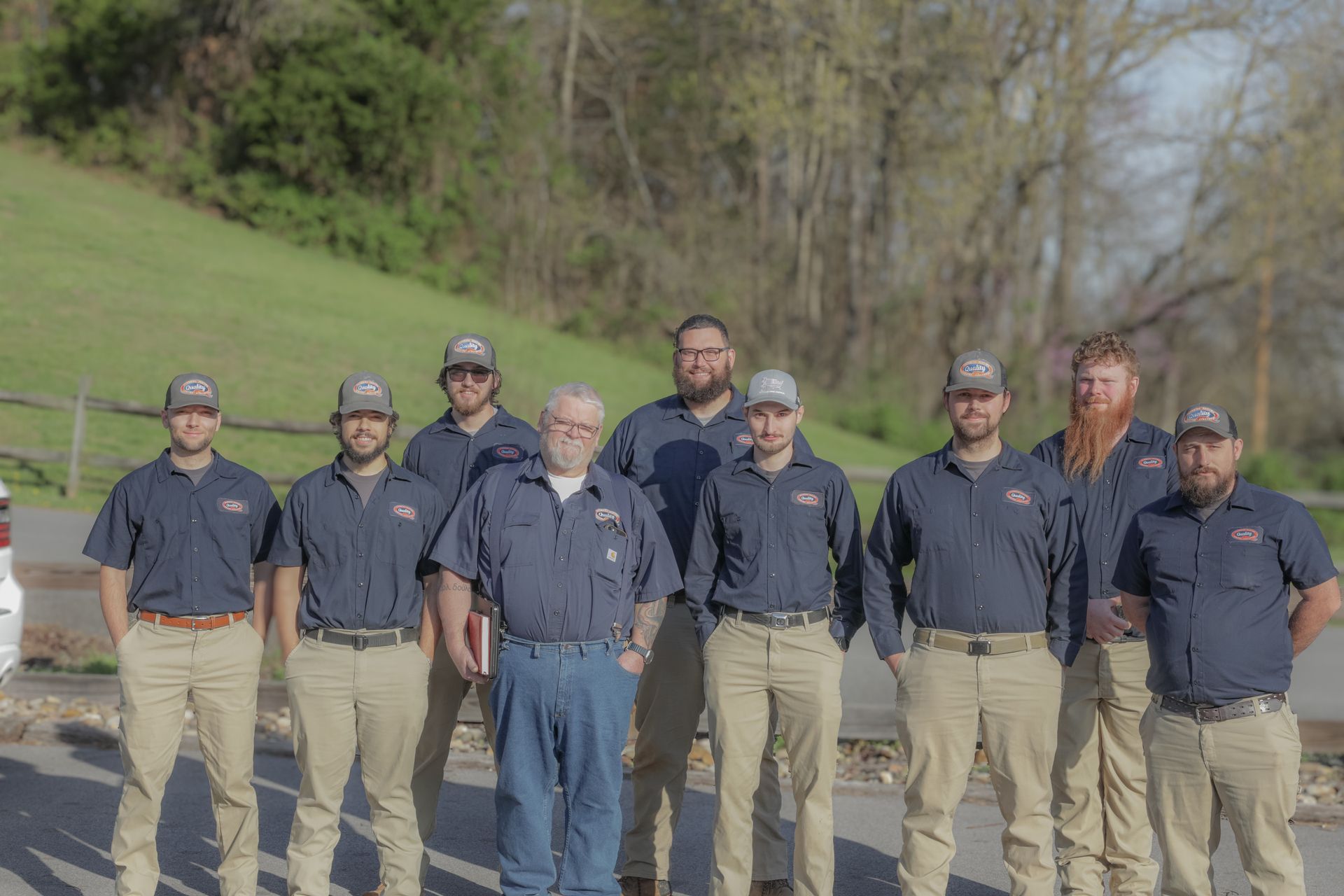 A group of nine people in uniform navy button-down shirts and khaki pants standing in a row outdoors on a sunny day.