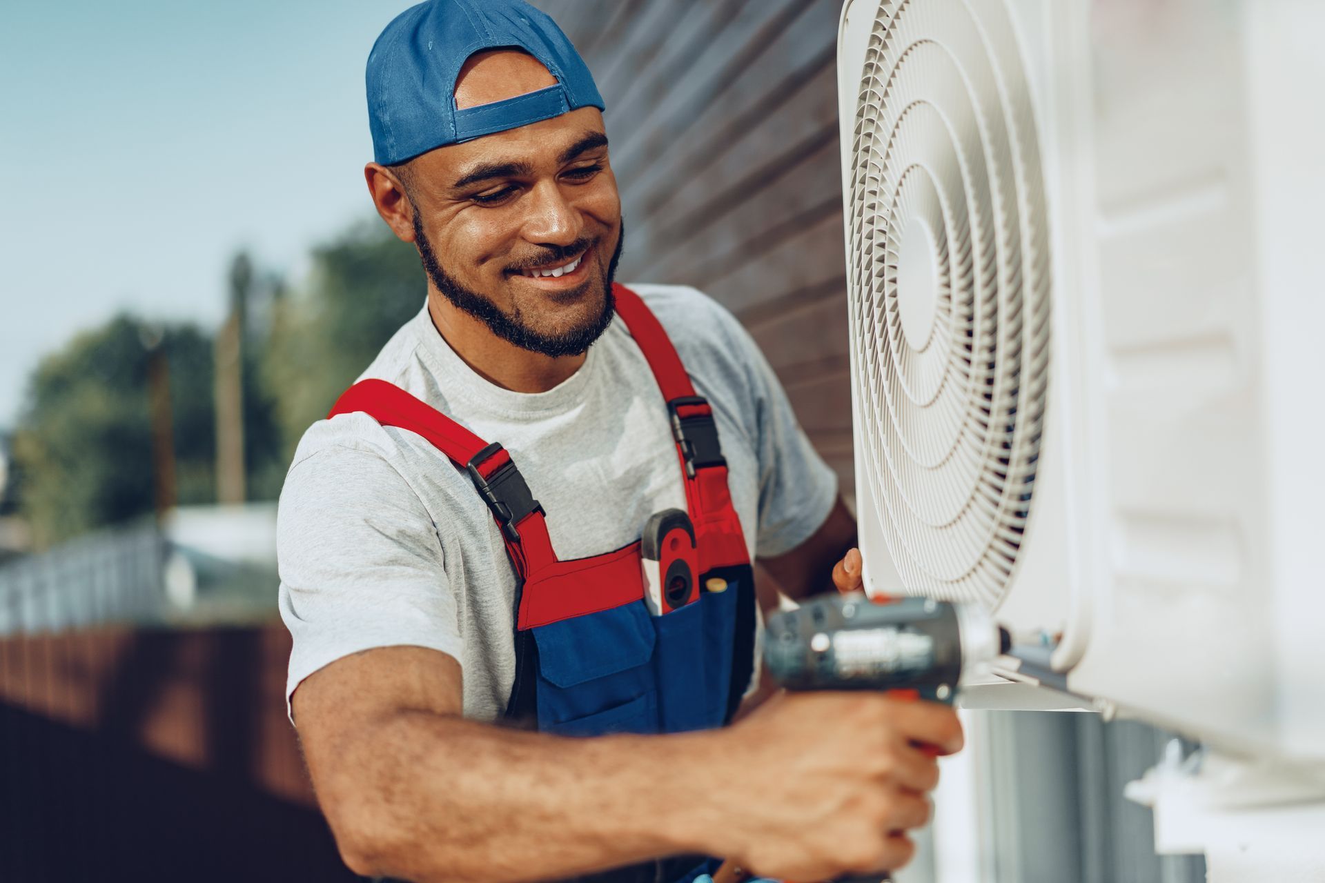 A person in a blue cap and work overalls uses a power drill to install an outdoor air conditioning unit.