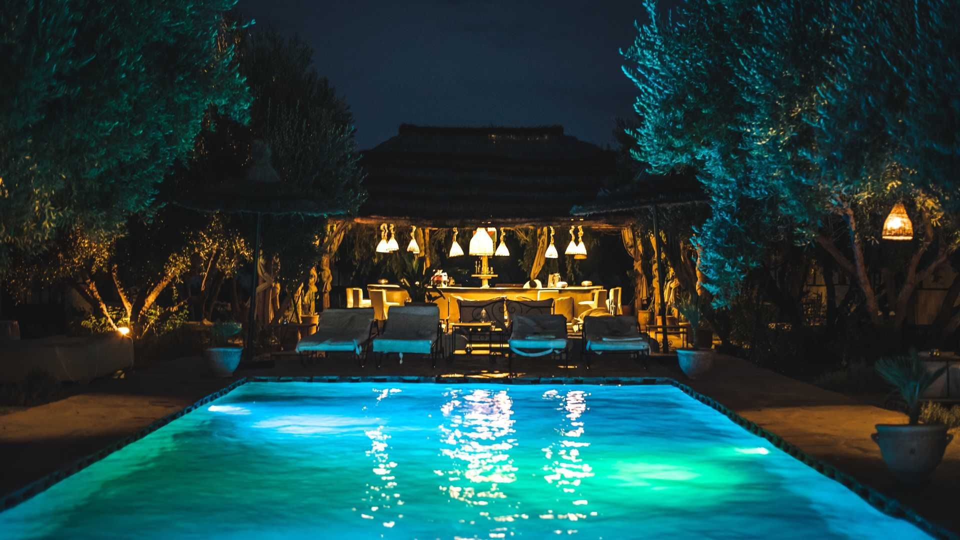 A large swimming pool is lit up at night with a gazebo in the background.