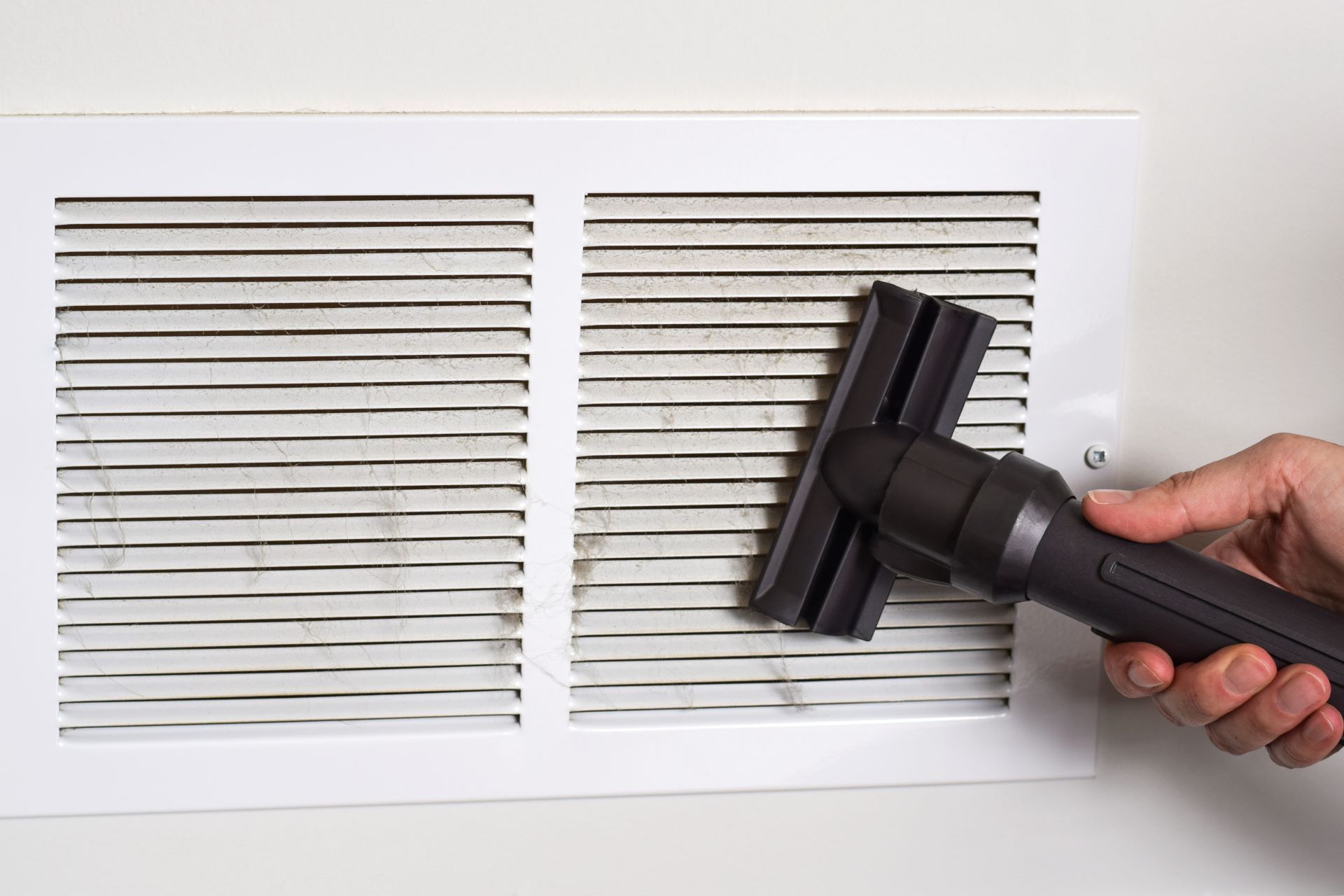 A hand uses a vacuum attachment to clean dust from the slats of a white wall-mounted air vent.