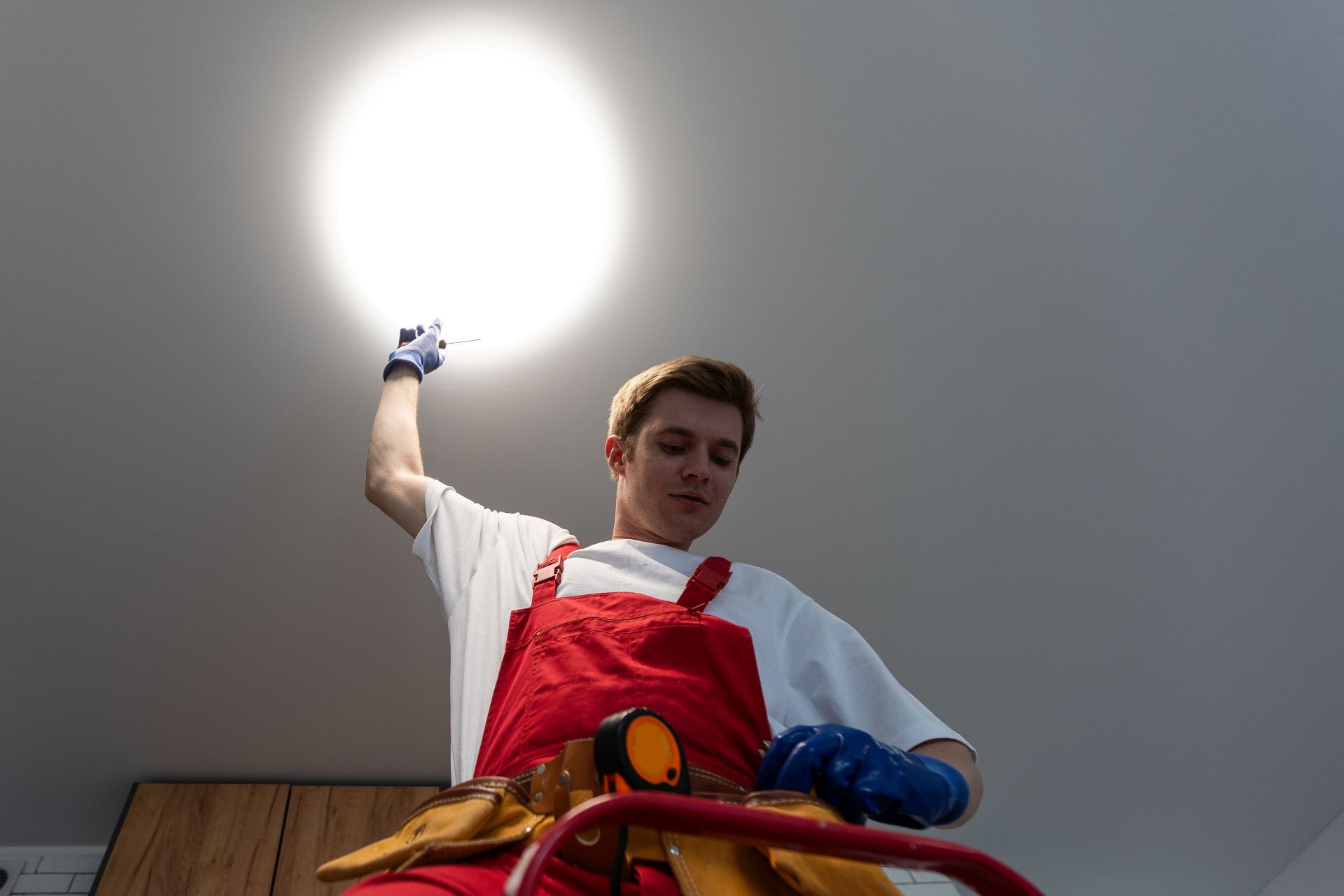 Man in red overalls on a ladder, working on a ceiling light.