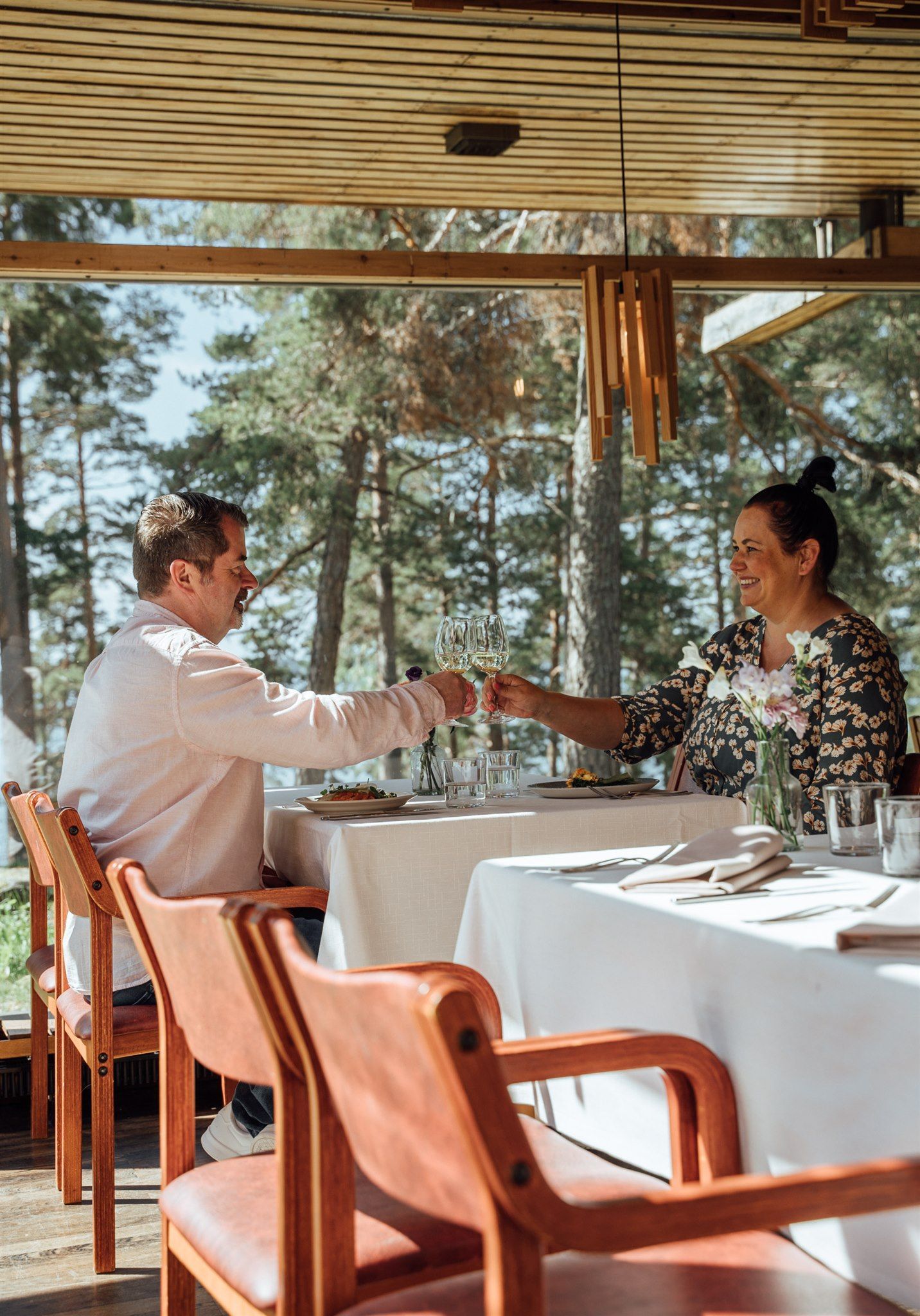 A person in a light shirt and a person in a patterned top toast with glasses of wine at an outdoor restaurant table.