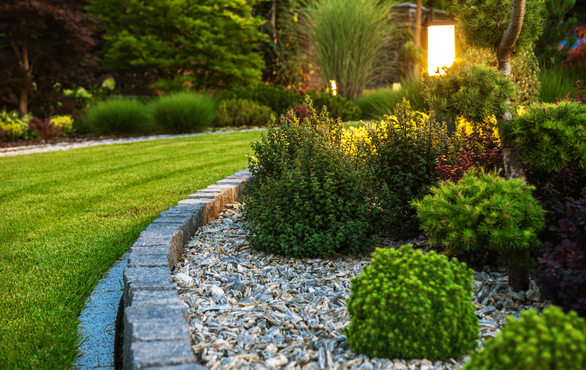 Green lawn and garden bed with various shrubs, bordered by stone bricks.