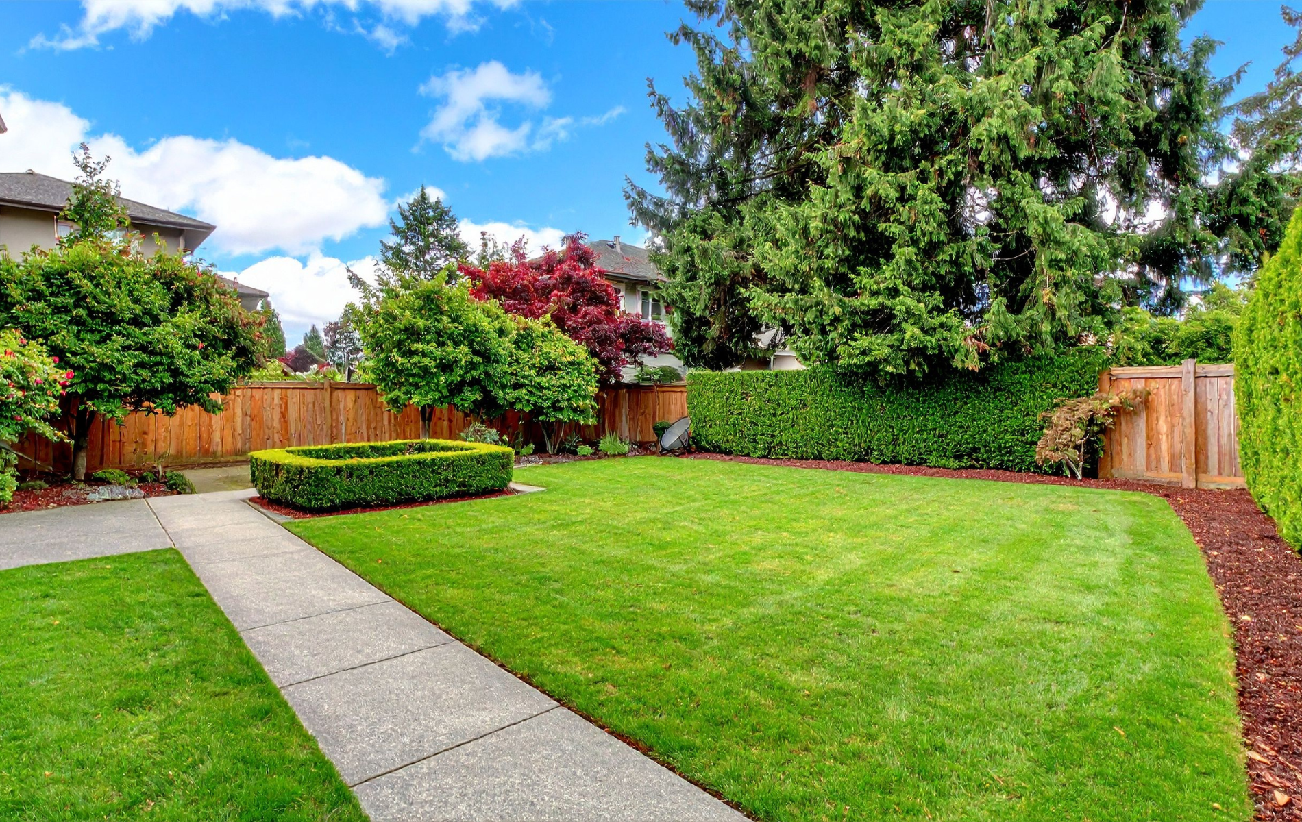 Well-maintained backyard with green grass, concrete path, wooden fence, and blue sky.