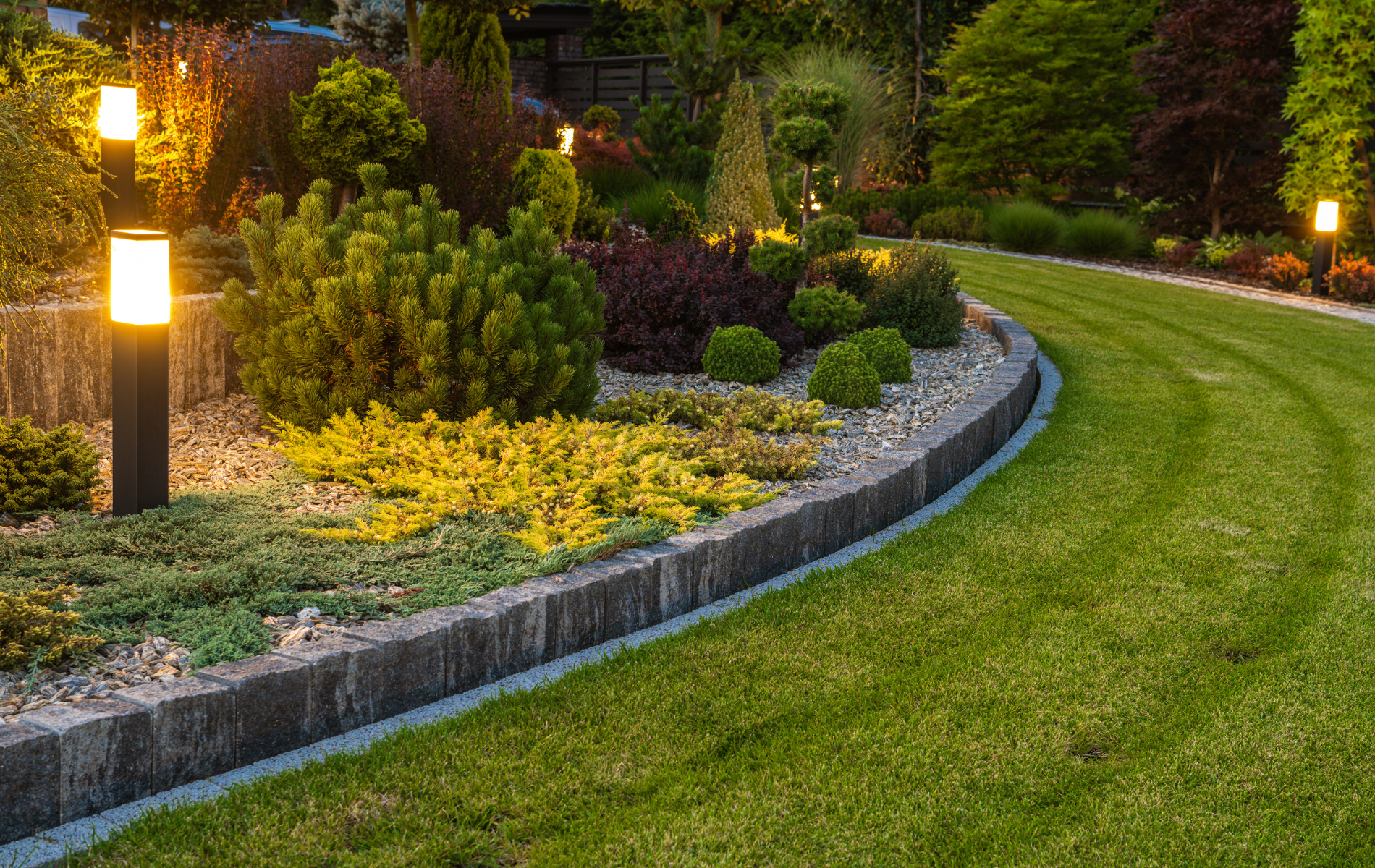 Lush garden bed with various plants, bordered by stone and lit by path lights, beside a green lawn.
