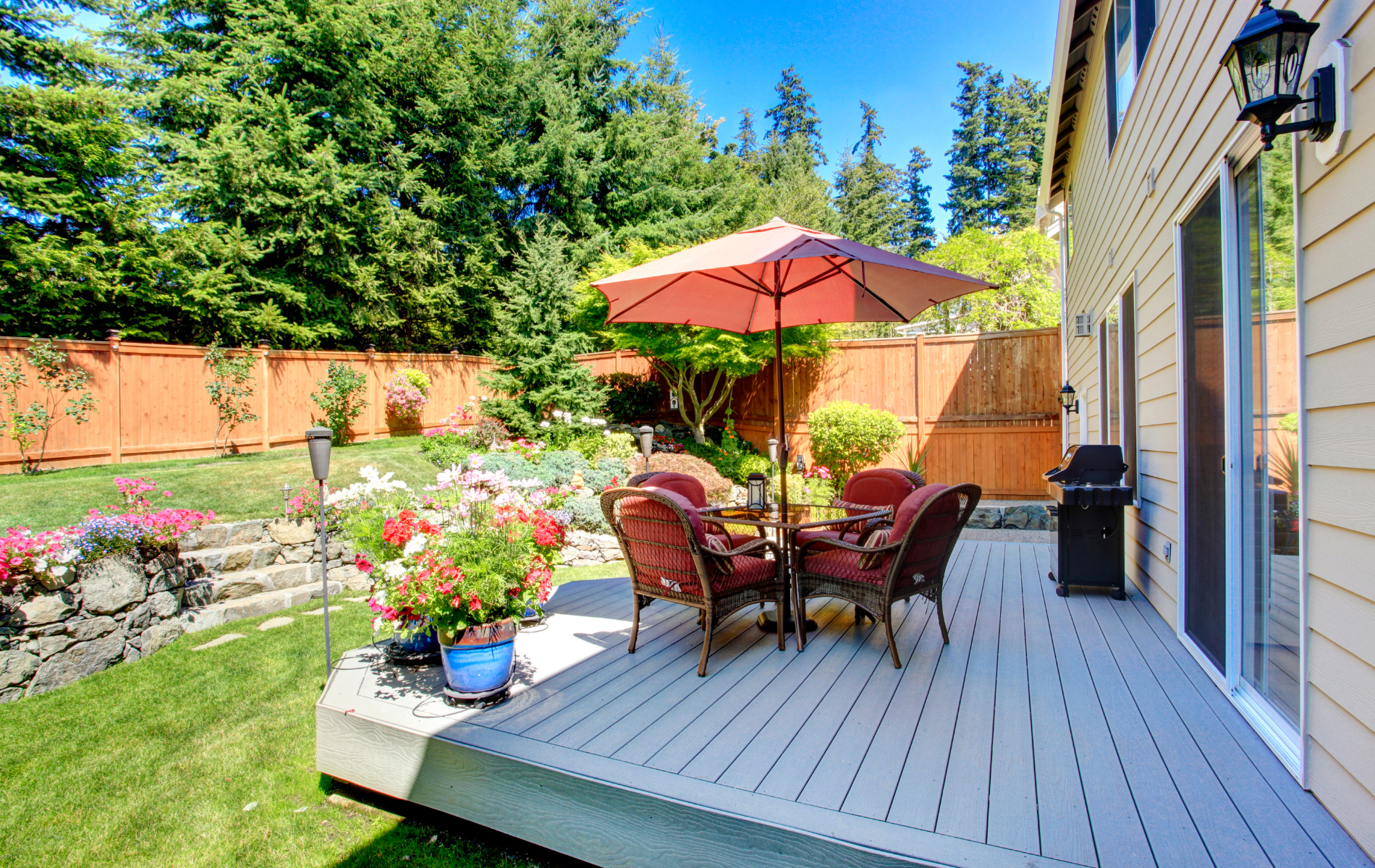 Backyard deck with patio furniture, umbrella, grill, and colorful flowers; tall trees and fence in the background.
